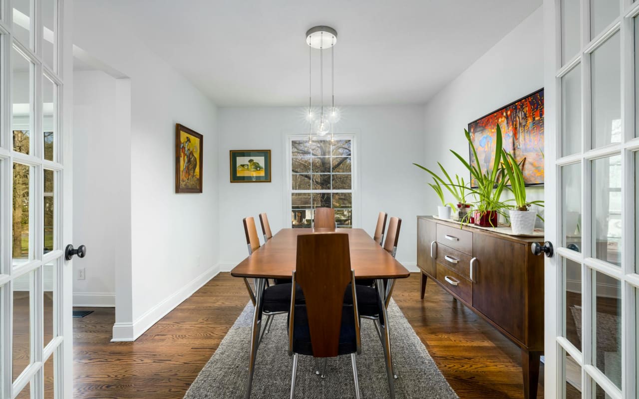 A dining room with a wooden table and chairs, a chandelier, a credenza on the left, and a doorway with two white doors.