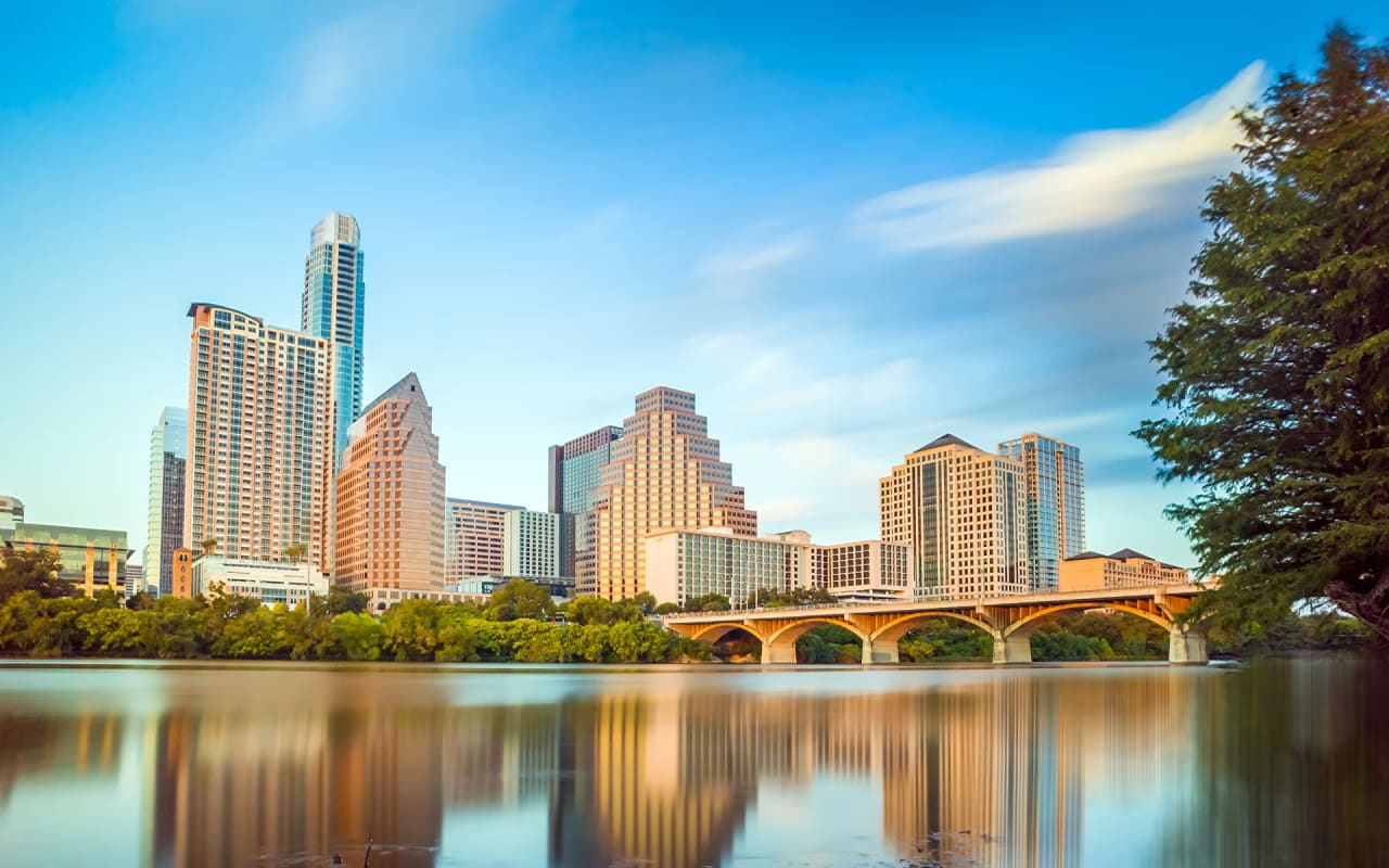 view of Austin, TX skyline from across a glassy river with a bridge in the foreground