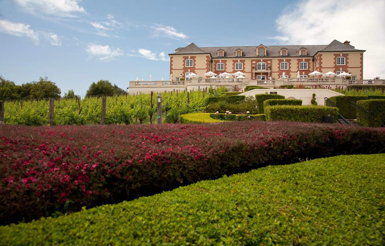 A large building with a tiled roof and arched windows. There is a vineyard and gardens in front of the building.