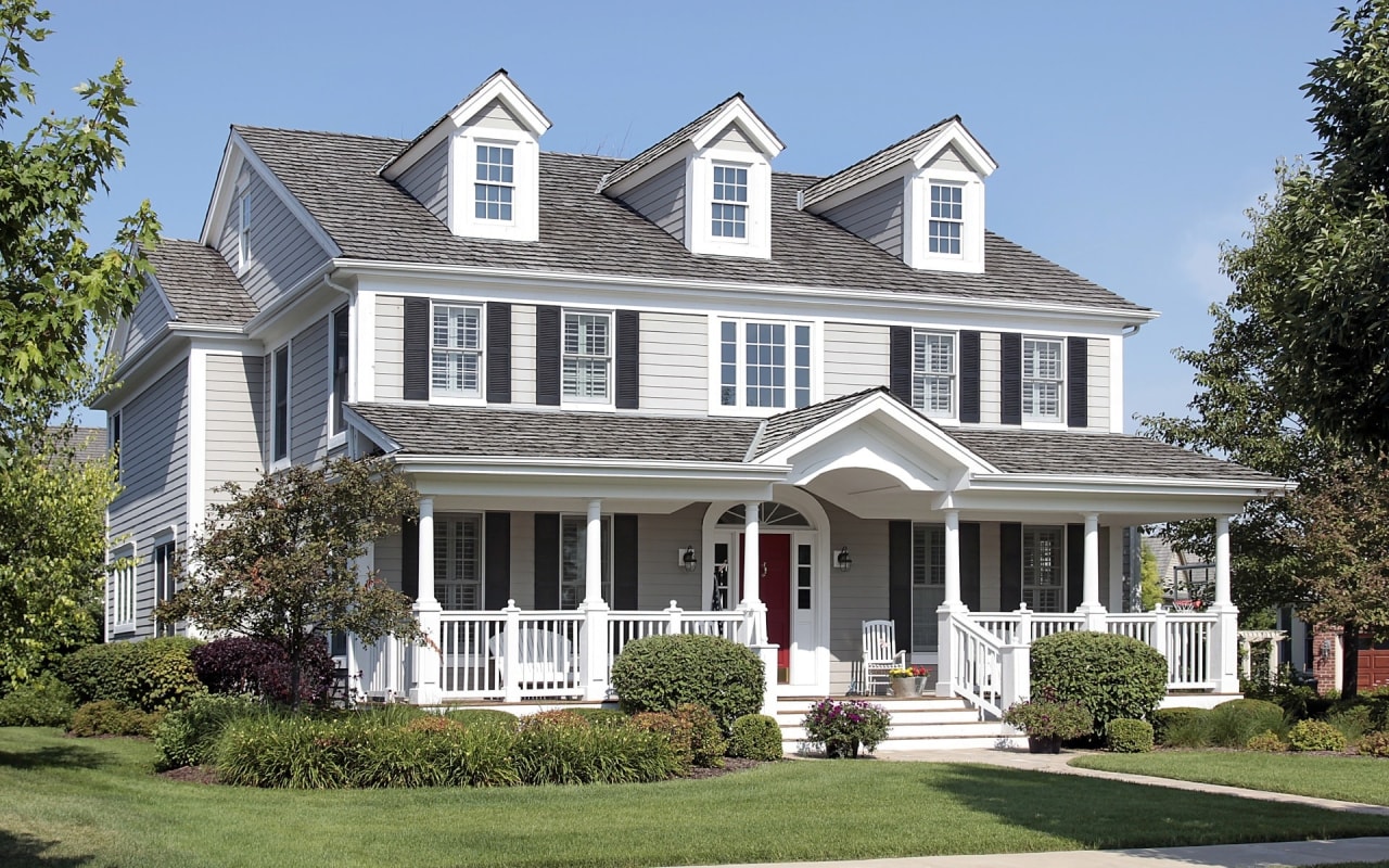 A large house with a pathway leading to the wraparound porch, surrounded by trees, bushes, and a green lawn.