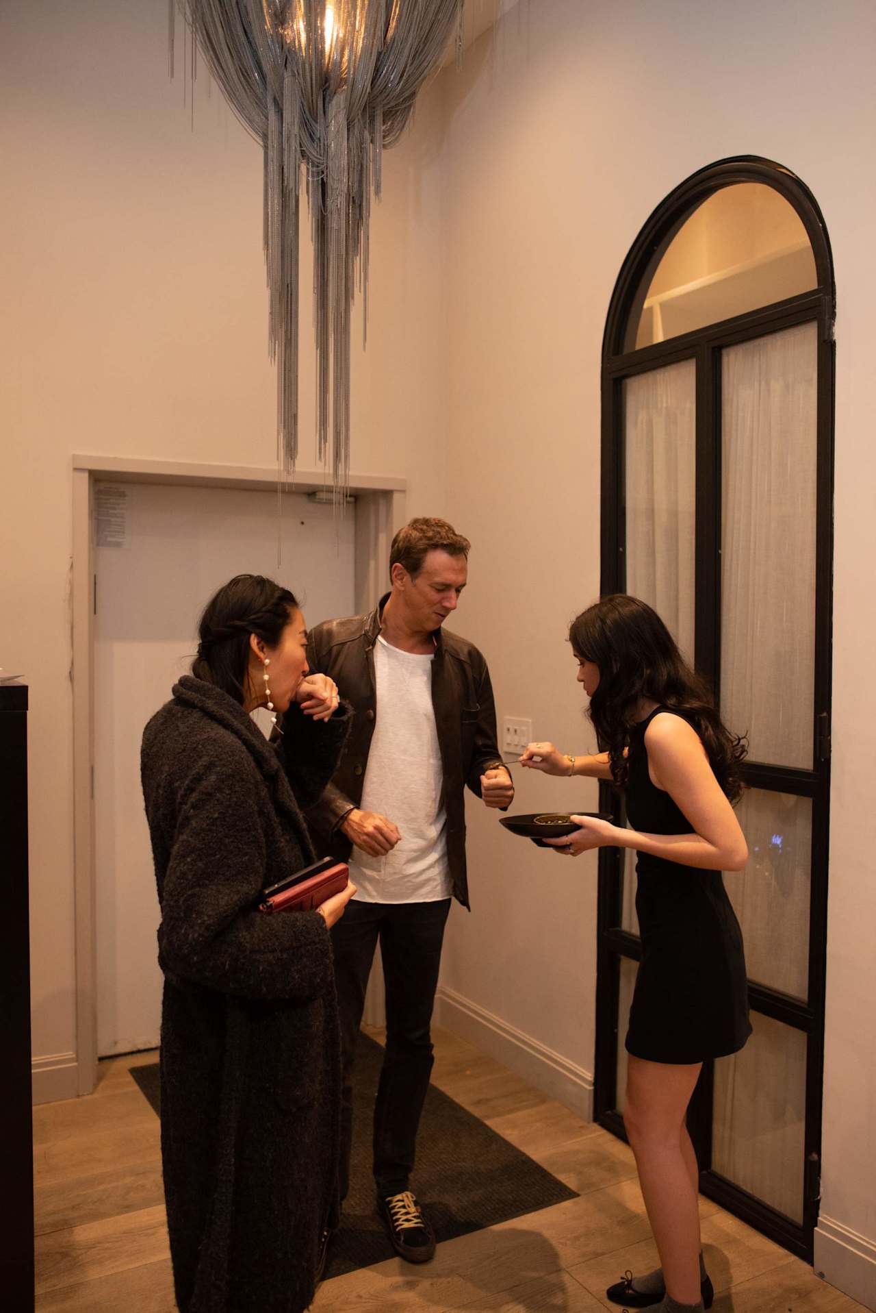 Three guests mingle beneath a chandelier as a woman serves food beside an arched window.