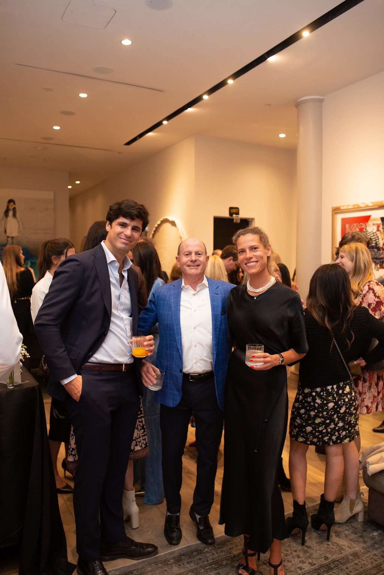 Three guests pose with drinks at a crowded indoor event.