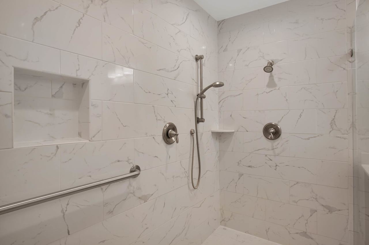 Close up of the inside of the ultra modern, extra large walk in shower featuring multiple shower heads, built in shelves, an assistance real, and white tile with light gray marbling at 1228 Winifred Drive, Tallahassee, Florida 32308