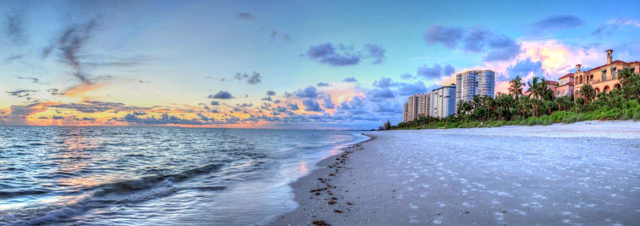 Beachfront with gentle waves, wide sandy shore, and palm-lined condominium buildings at sunset.