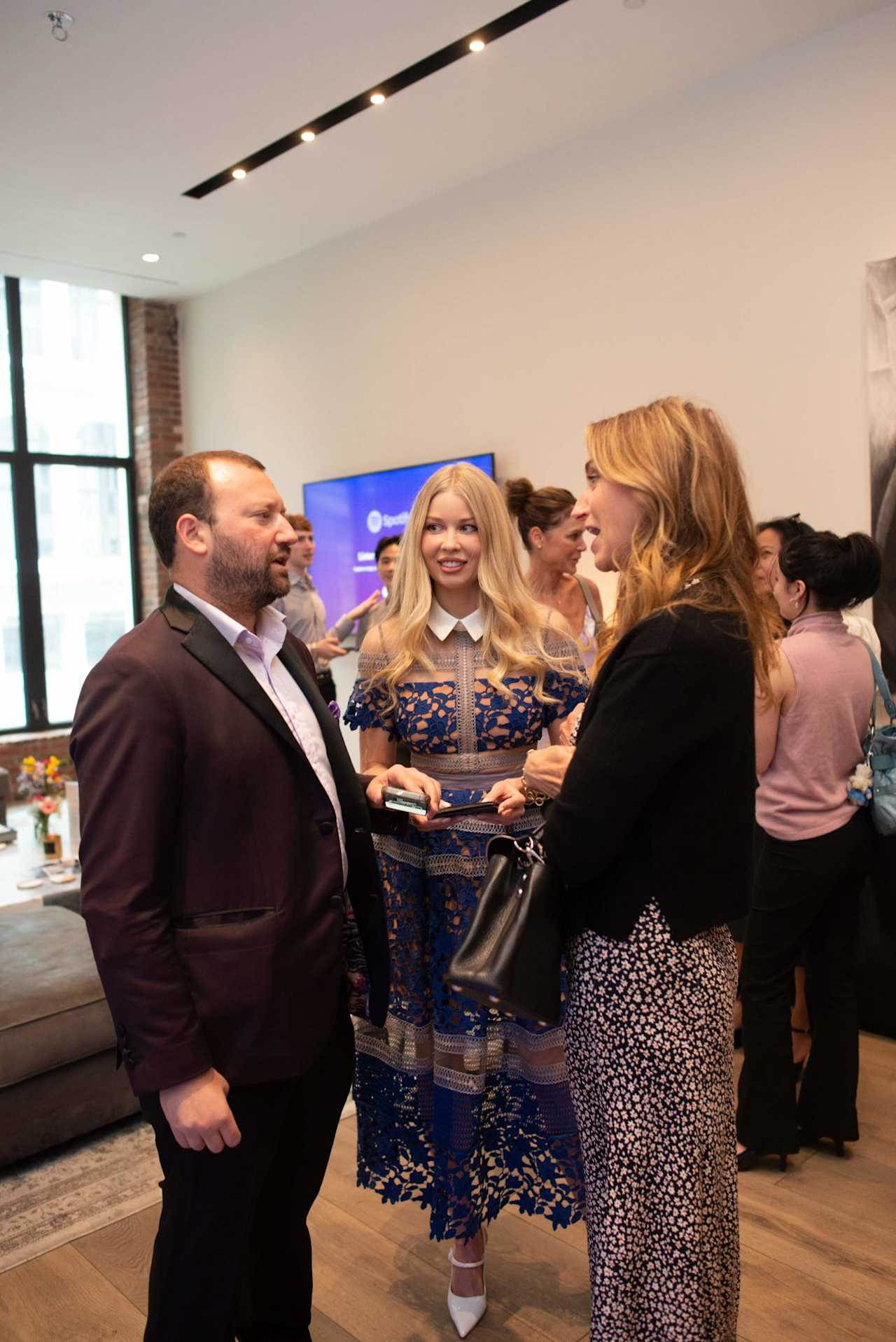 Guests mingle at a real estate gathering, including a woman in a blue lace dress.