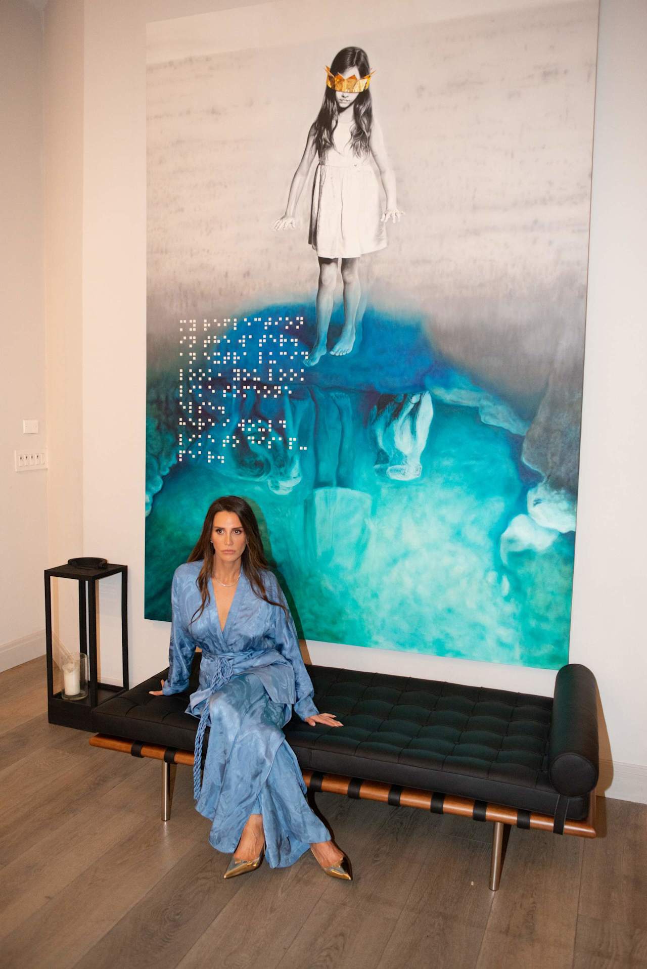Woman in a blue patterned outfit seated before a large turquoise artwork.