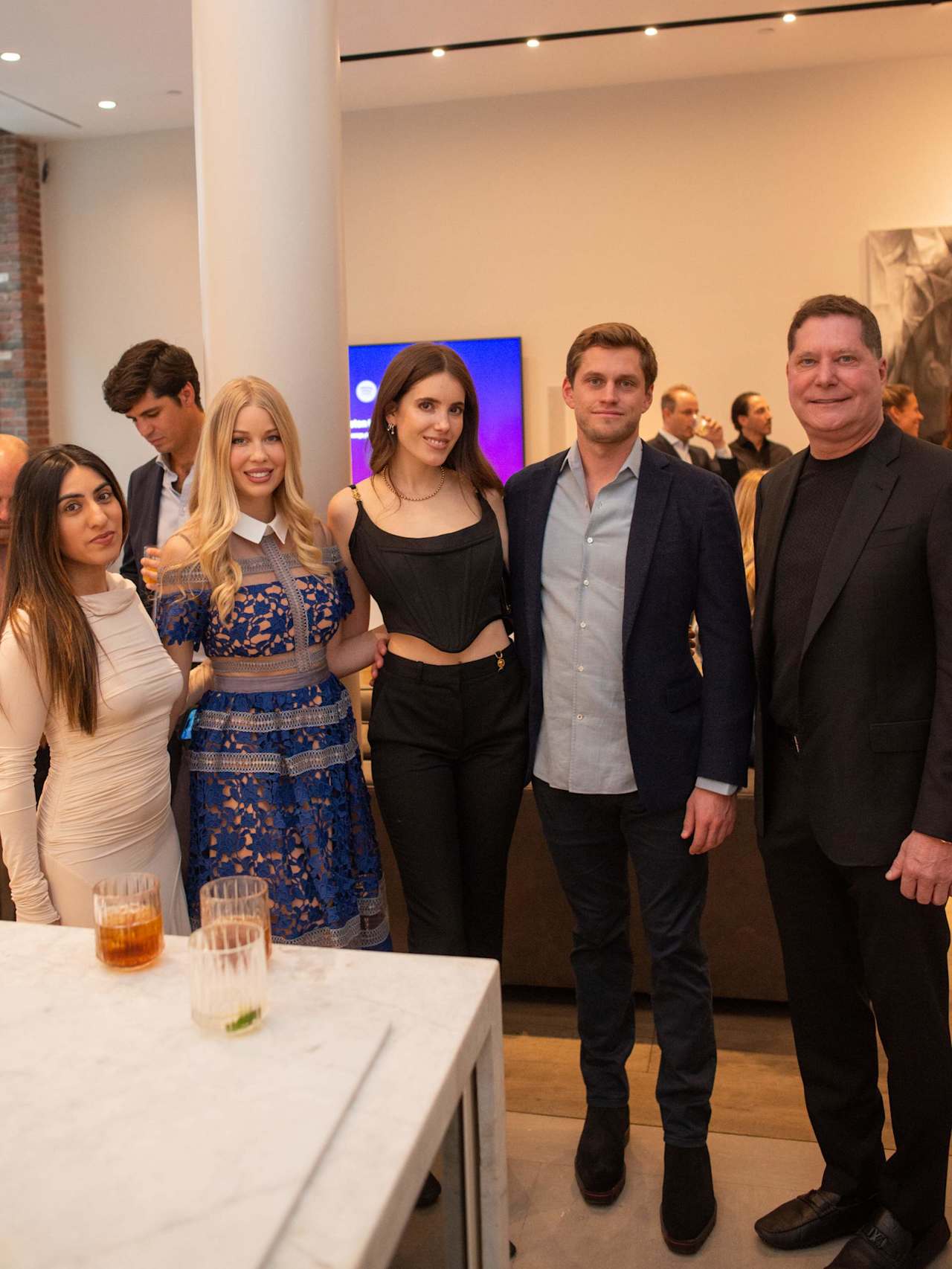 Adults pose together at a gathering, including a woman in a blue lace dress and a man in a navy blazer.