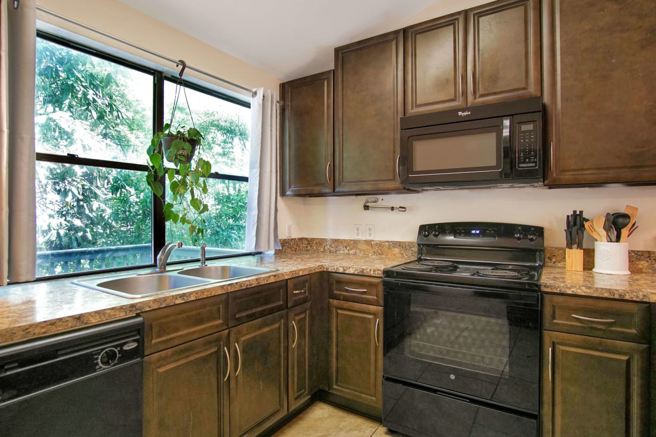 A kitchen with dark cabinets and modern appliances. The kitchen has a large window above the sink, allowing natural light to fill the space. The countertops are light-colored, and the floor is tiled.