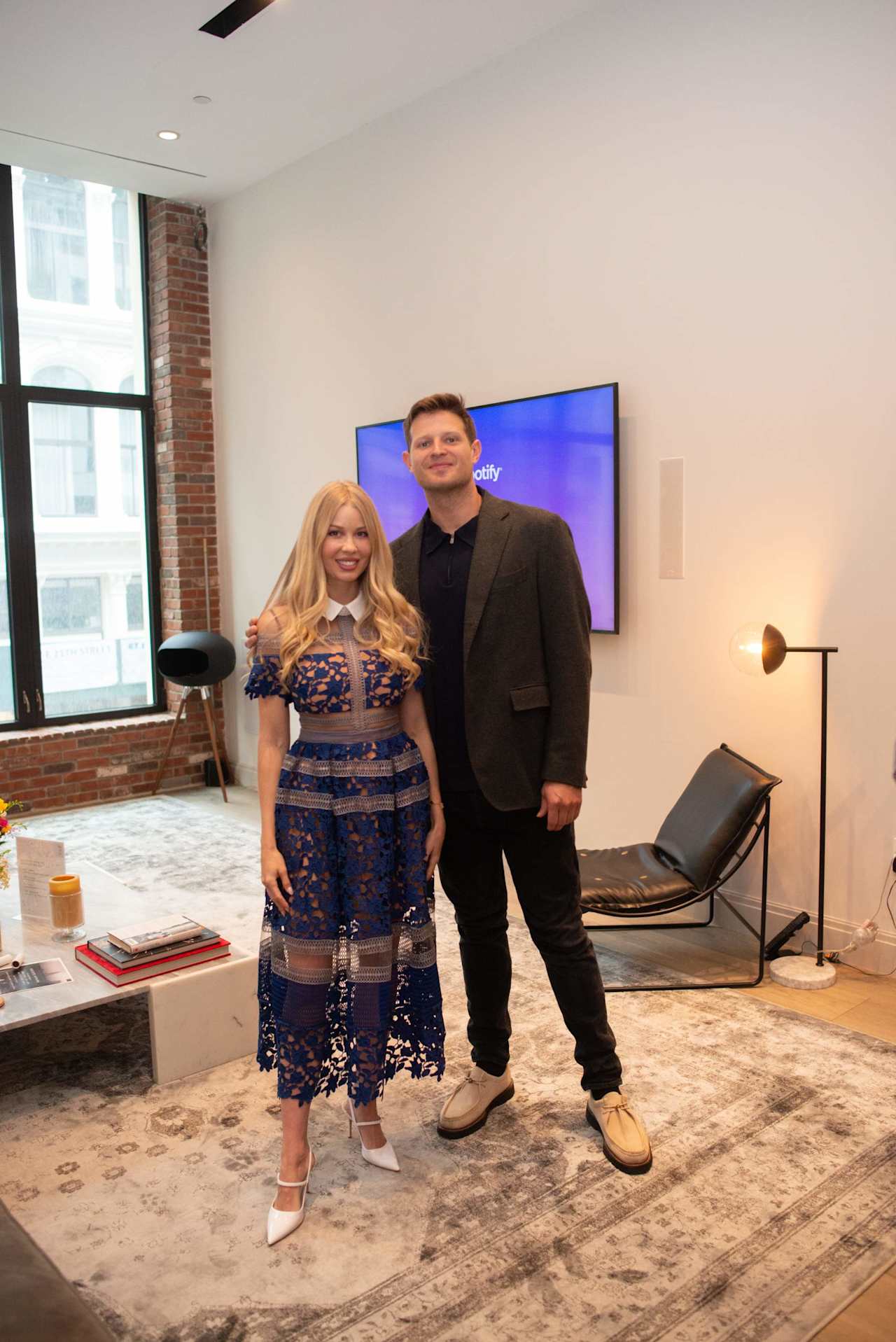 A woman in a blue lace dress and a man in a dark blazer pose together in an office lounge.