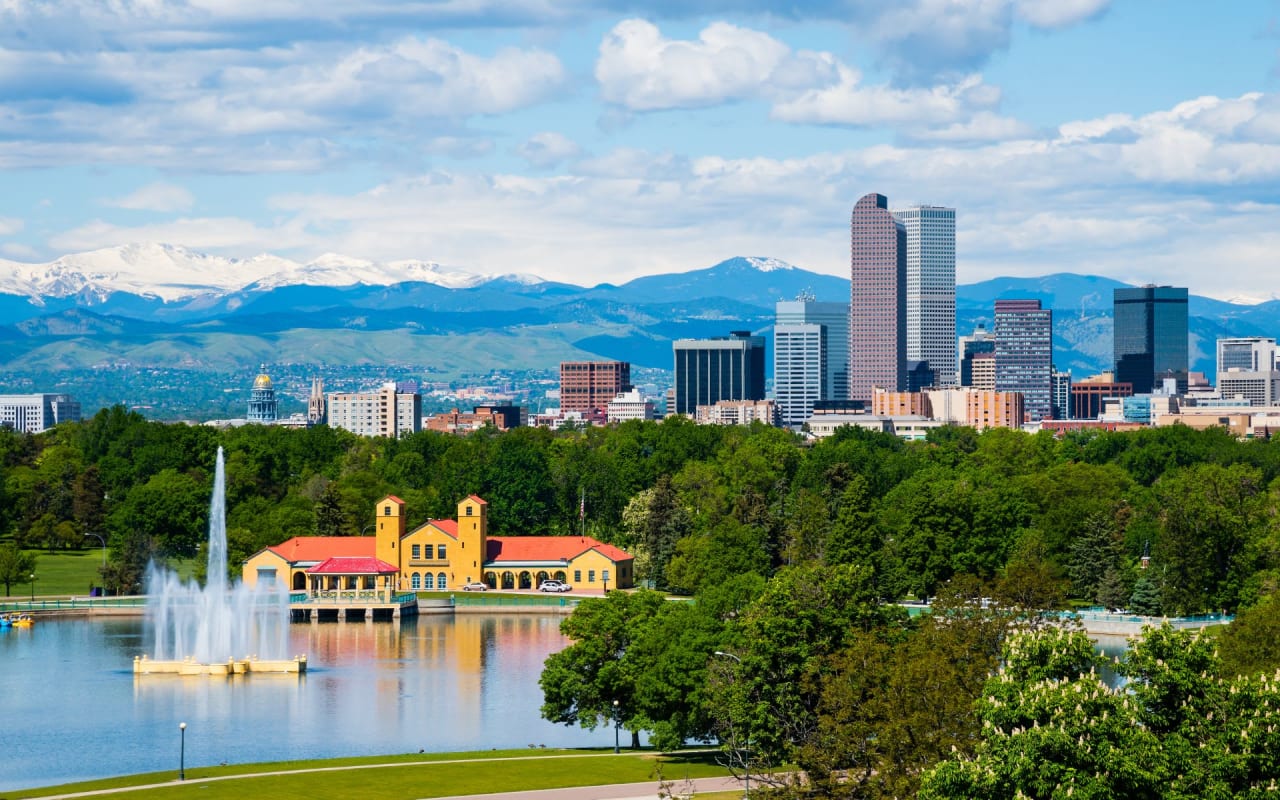 Denver City Park's beautiful fountain with city skyline and mountain views amid lush greenery.