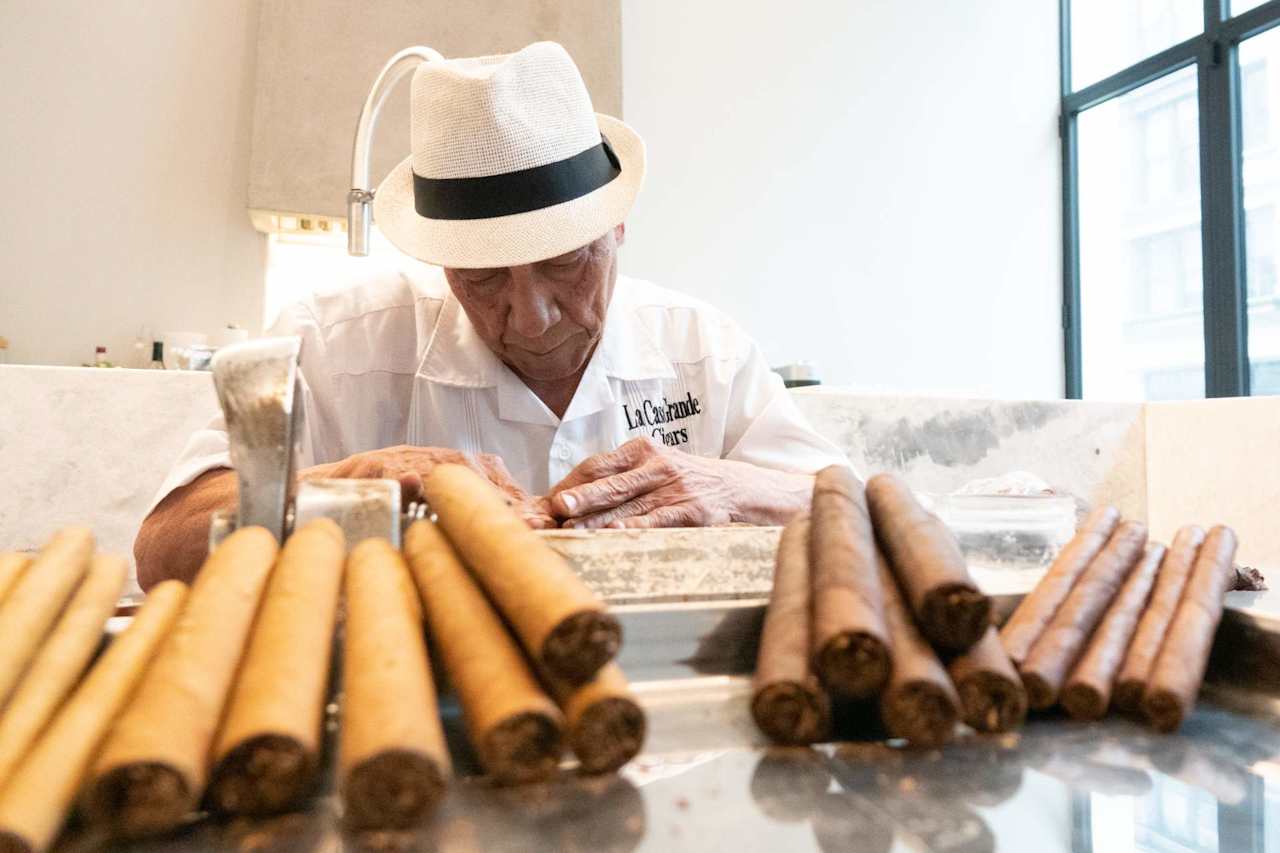 Cigar maker hand-rolls cigars behind rows of finished cigars.