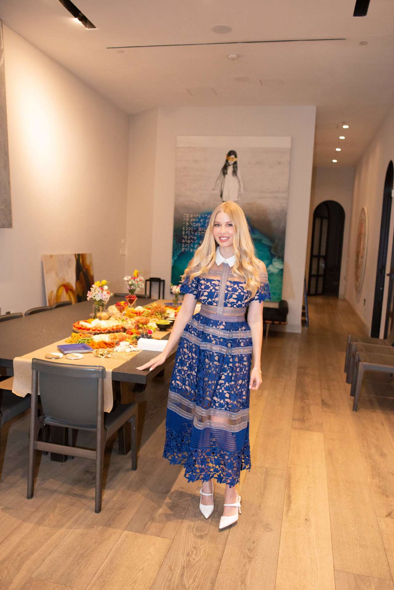 Blonde woman in a blue lace dress stands beside a table of food in an art-filled room.