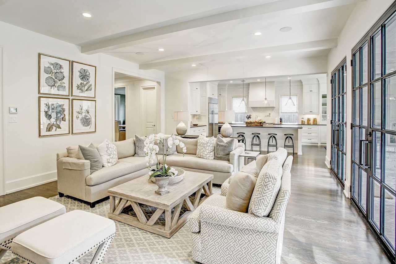 A modern living room with various furniture, a patterned rug, two windows on the left side, and a kitchen at the back.