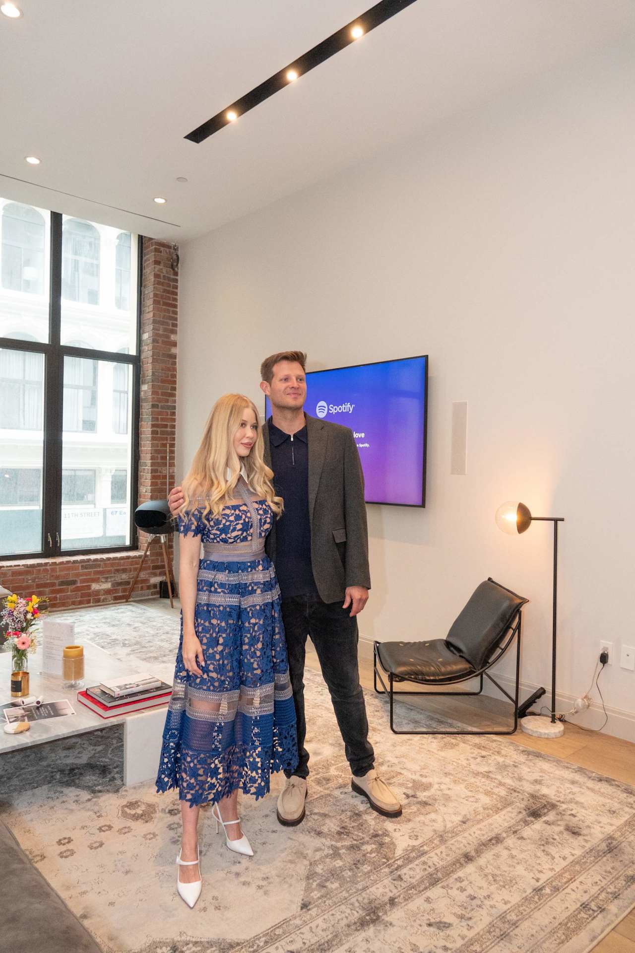 Two adults pose in an office lounge beside a screen displaying Spotify.