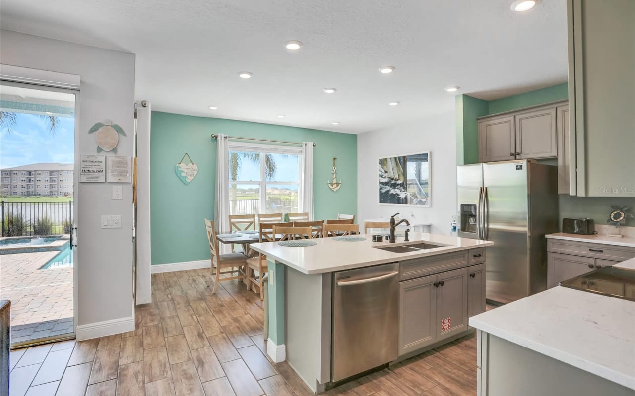 A kitchen with a stainless steel refrigerator, a sink with a faucet, a dining table, chairs, cabinets, and a large window.