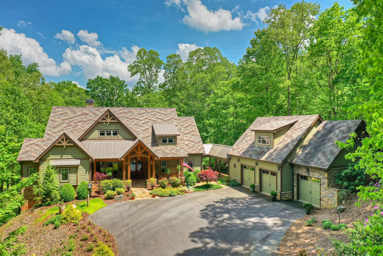 A large house with a driveway and two garages sits on top of a hill. Trees surround the house.