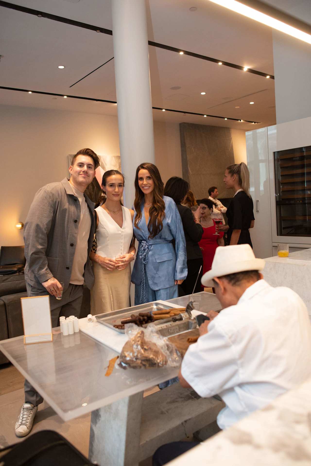 Three guests pose together at a gathering while other attendees mingle nearby.
