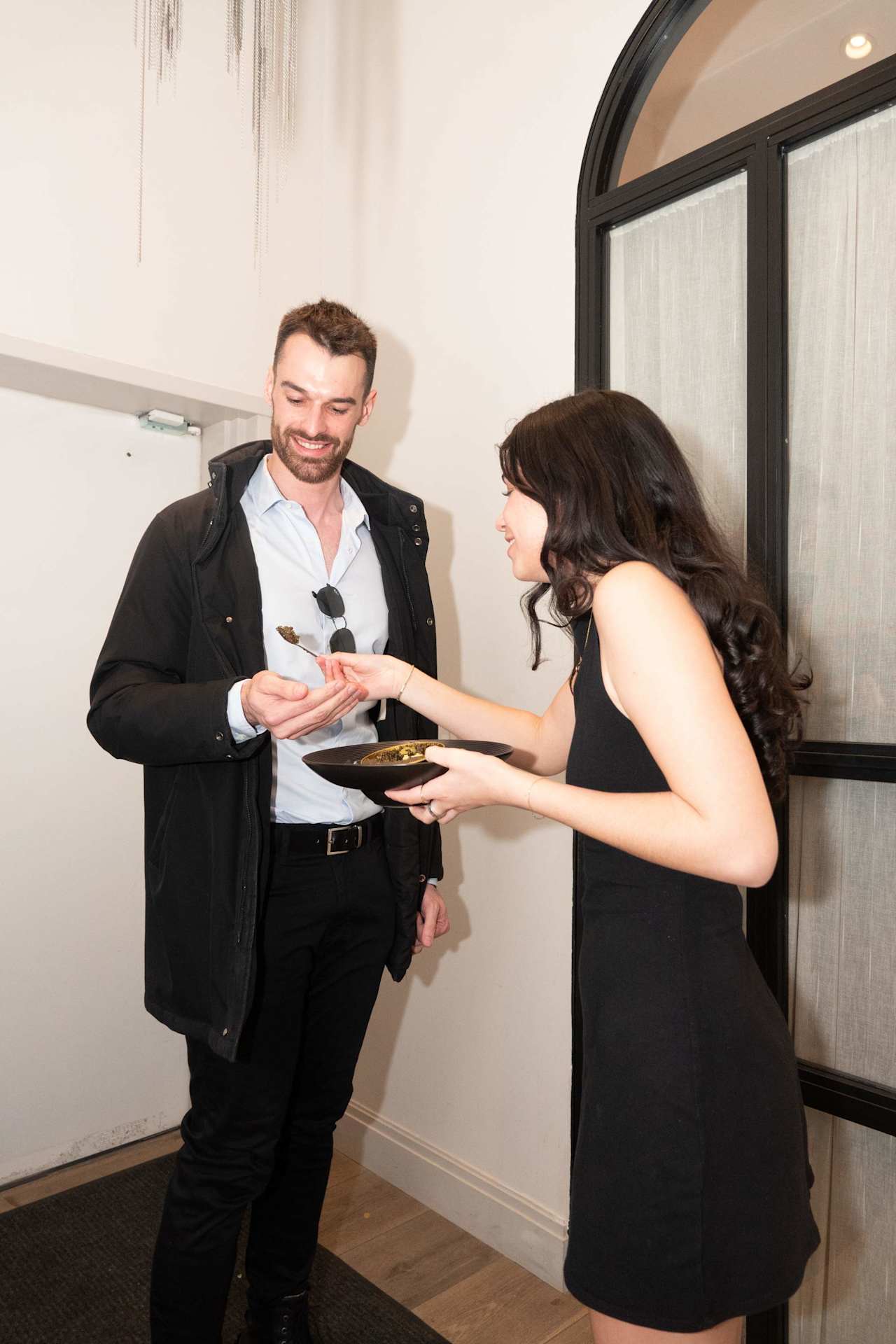 A woman offers a man a bite of food from a black bowl at an indoor event.