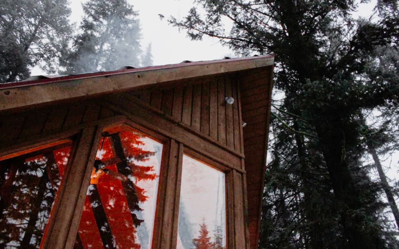 A rustic wooden cabin in a forest with several windows, a peaked roof,  and a chimney.