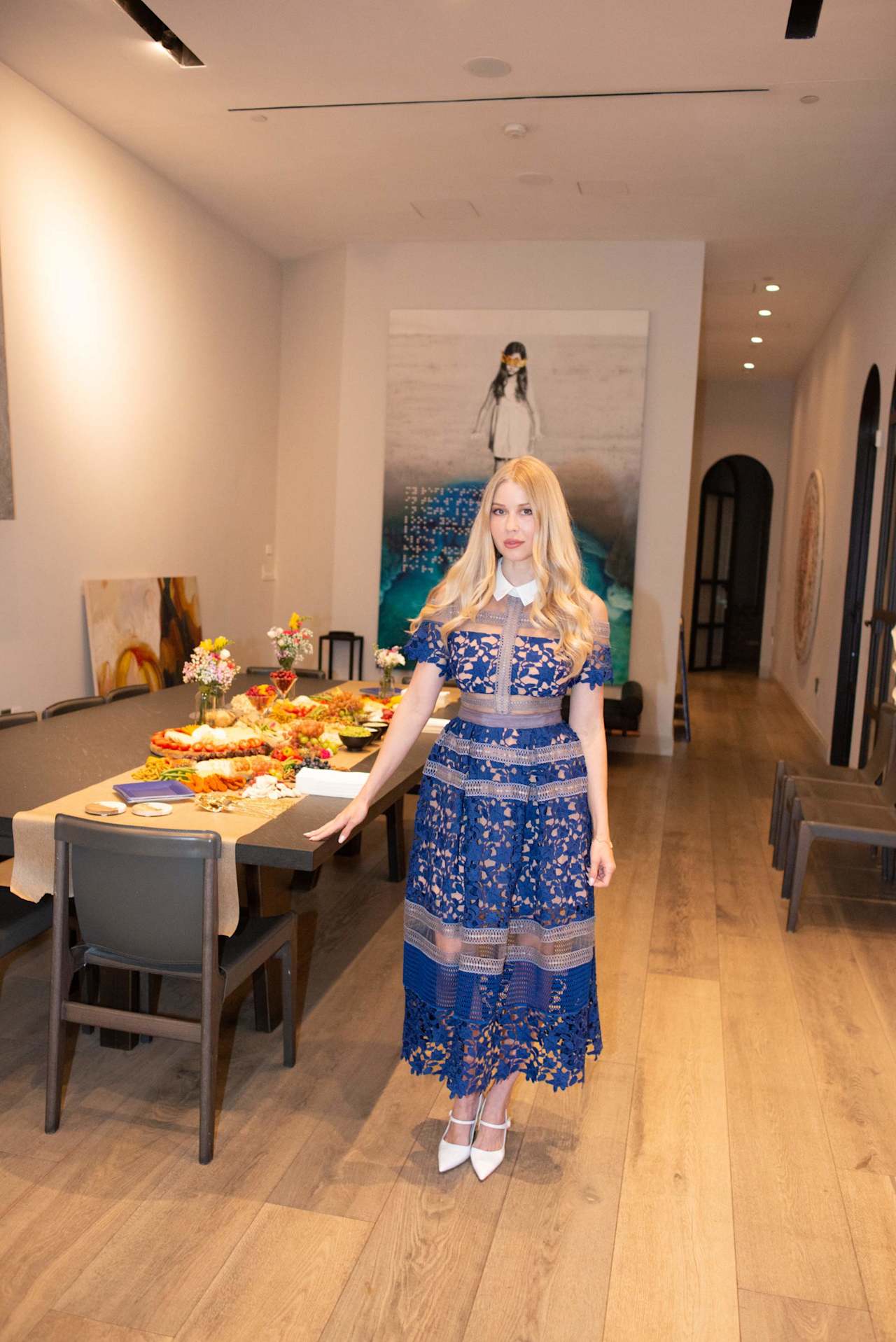 Woman in a blue lace dress stands beside a table laid with food and flowers.