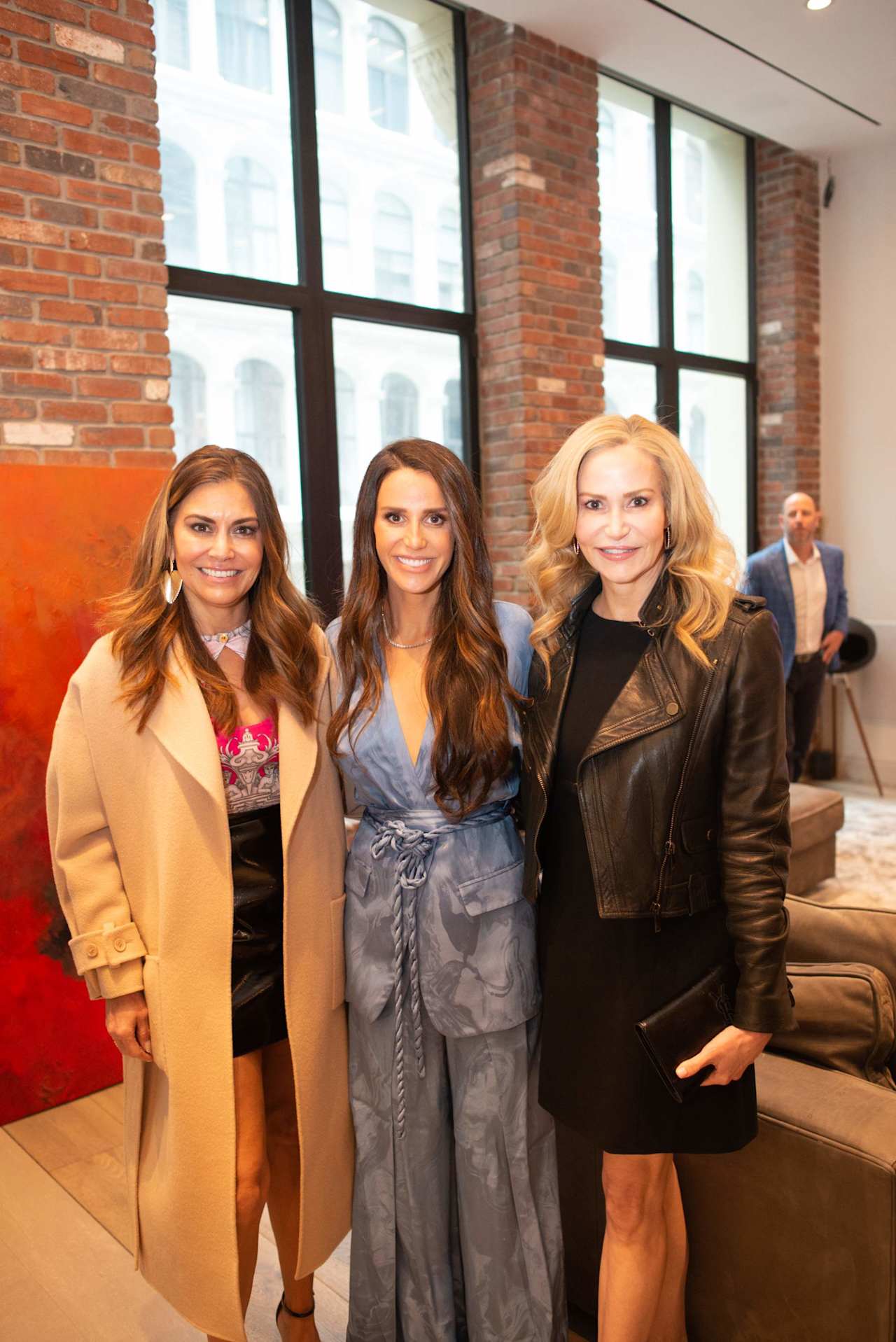 Three women pose together at an indoor gathering, with brick walls and large windows behind them.