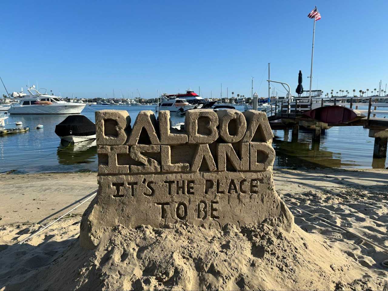 Sand sculpture reading 'Balboa Island: It's the Place to Be,' with boats in the background.