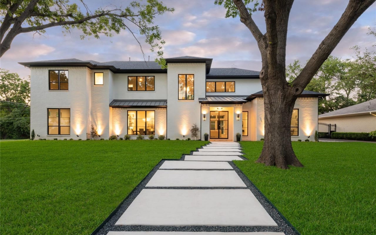 A white two-story house with large windows, a covered porch, a concrete walkway, and trees on the front lawn.