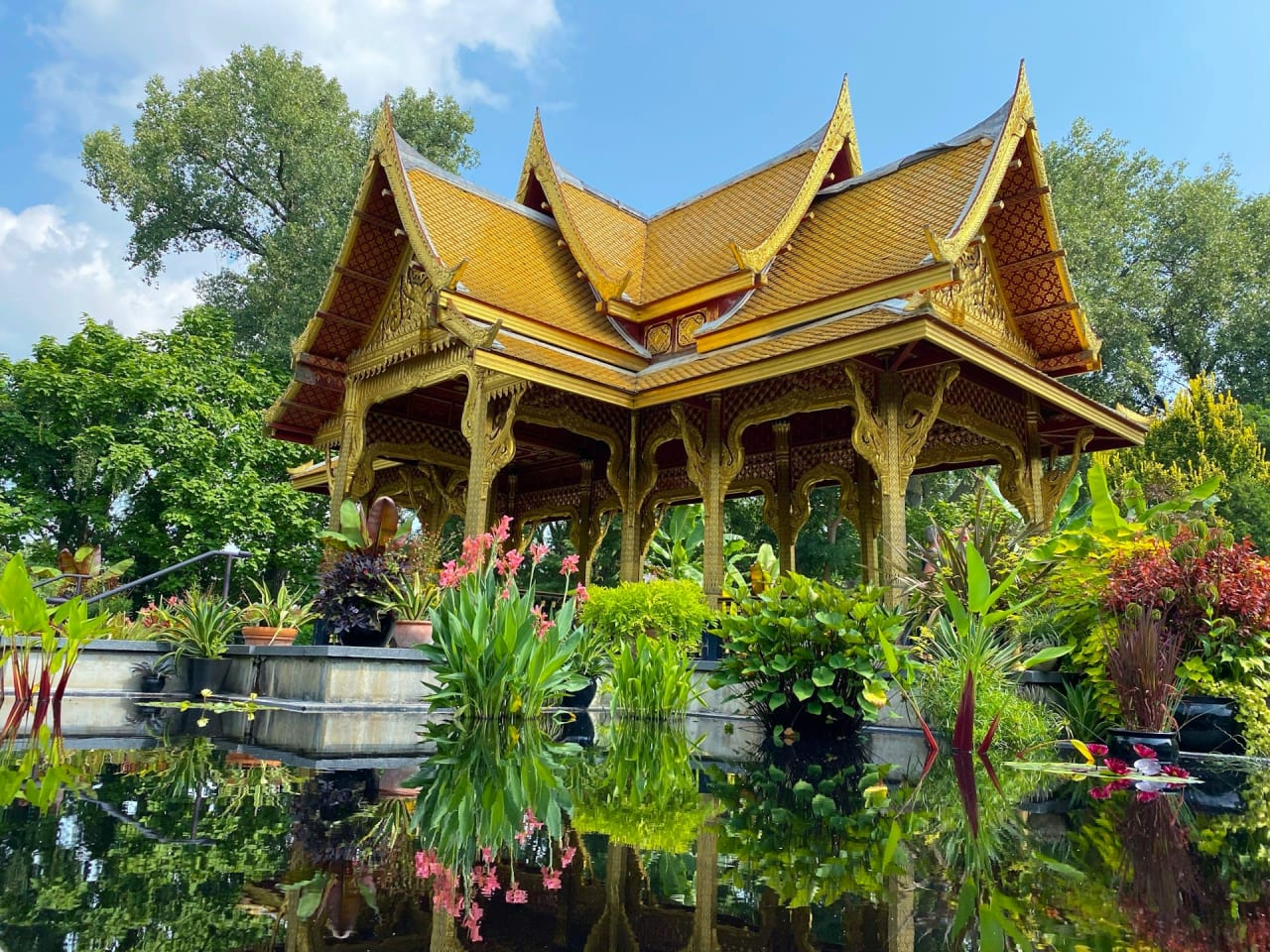 An ornate golden pavilion with a red roof sits on a pond in a botanical garden, surrounded by plants and trees.