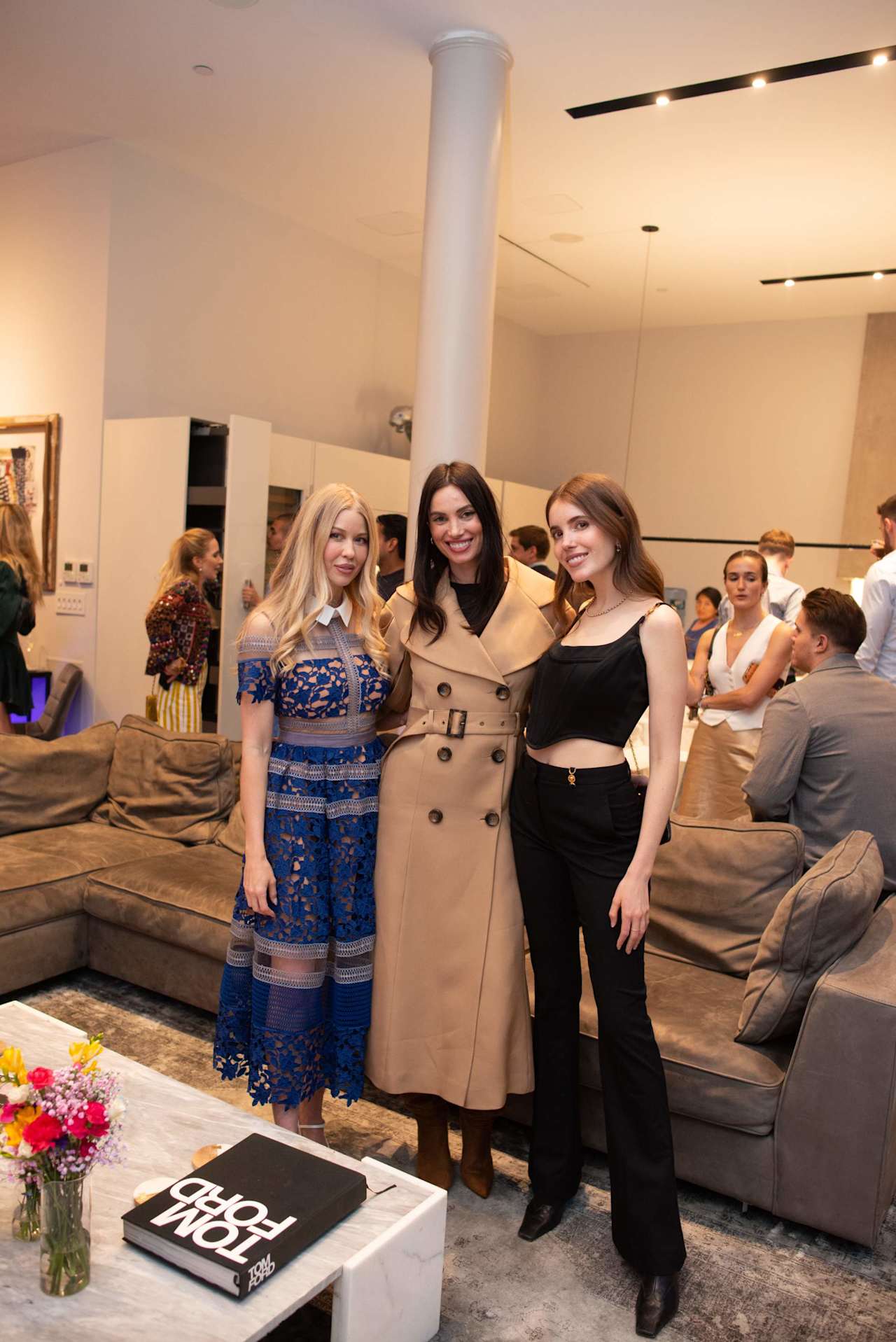 Three women pose at an indoor event beside brown lounge sofas.