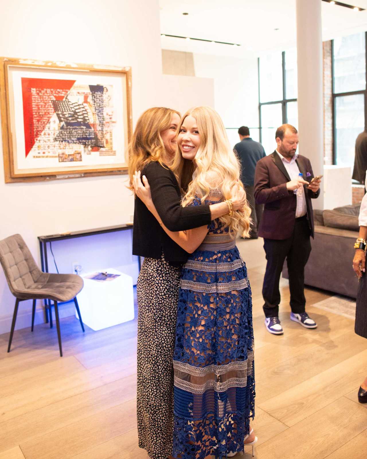 Two women embrace at an indoor gathering, one wearing a blue lace dress.