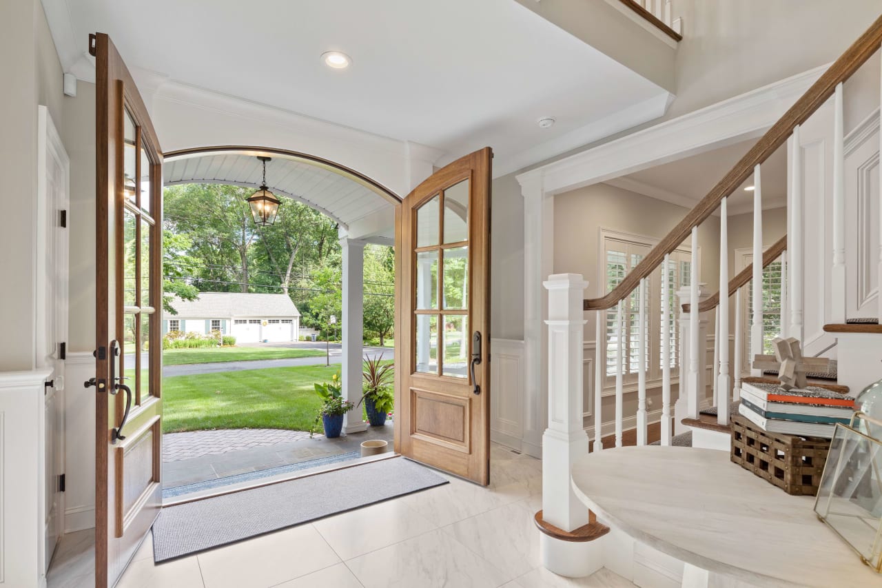 Bright and welcoming foyer with an open door, showcasing a view of the green lawn and a staircase with wooden railings.