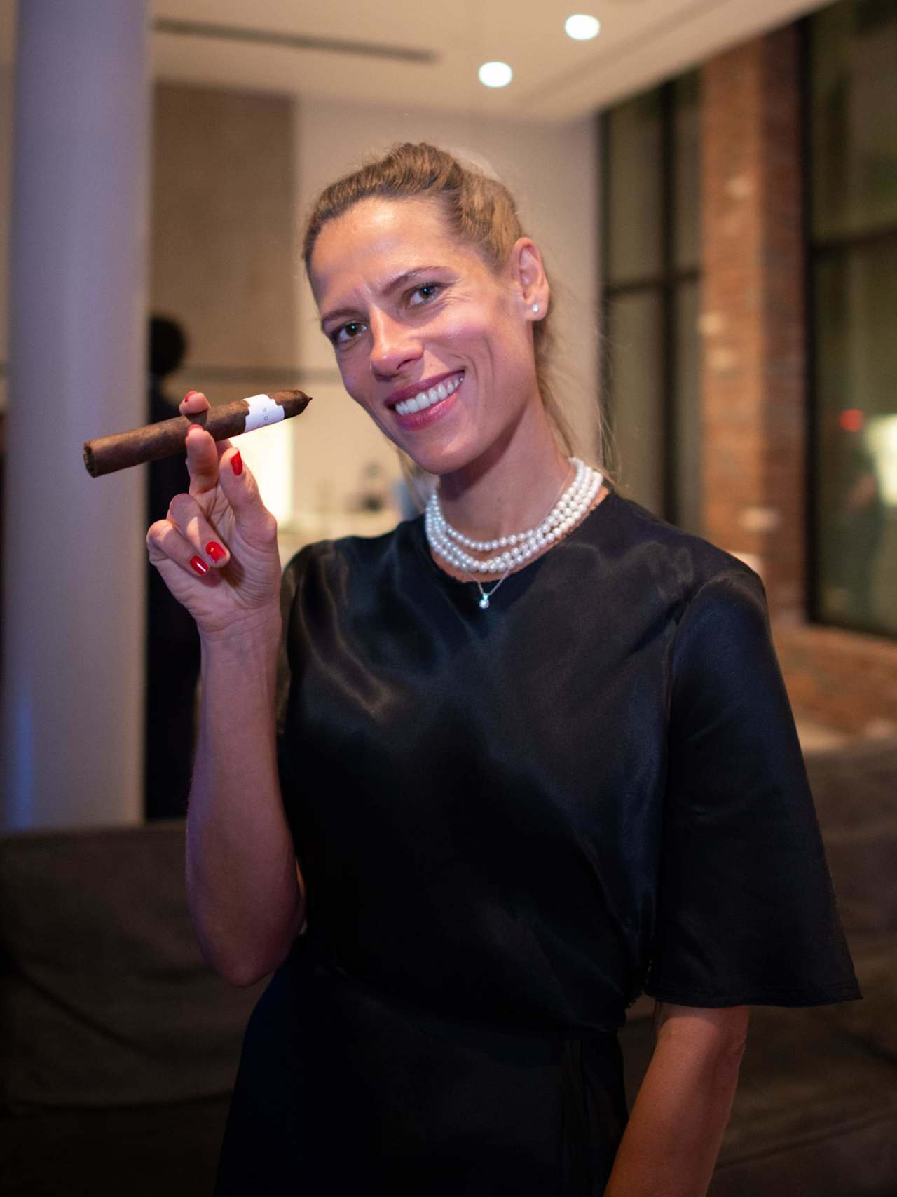 Smiling woman in a black top and pearl necklaces holding a cigar.