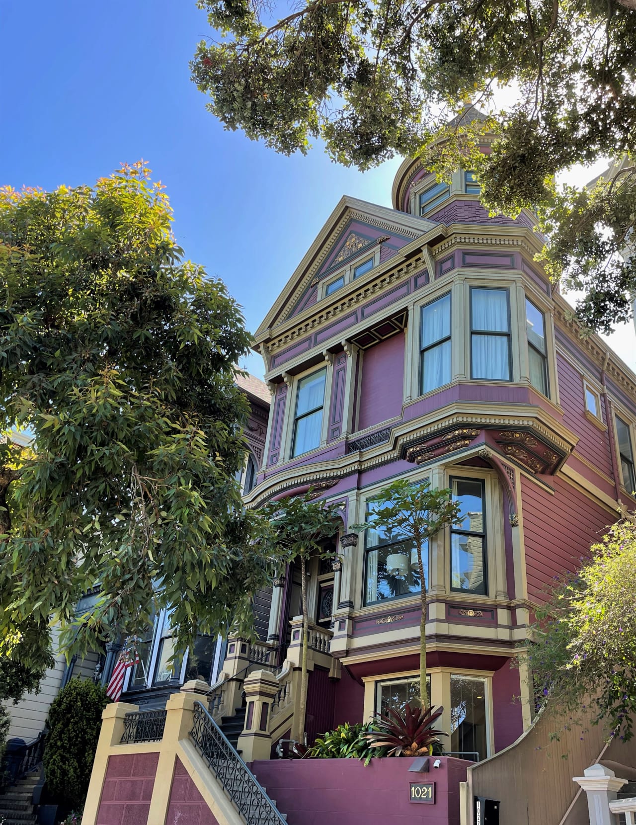 A purple and yellow Victorian house with stairs leading up to the front porch and is surrounded by tall trees.