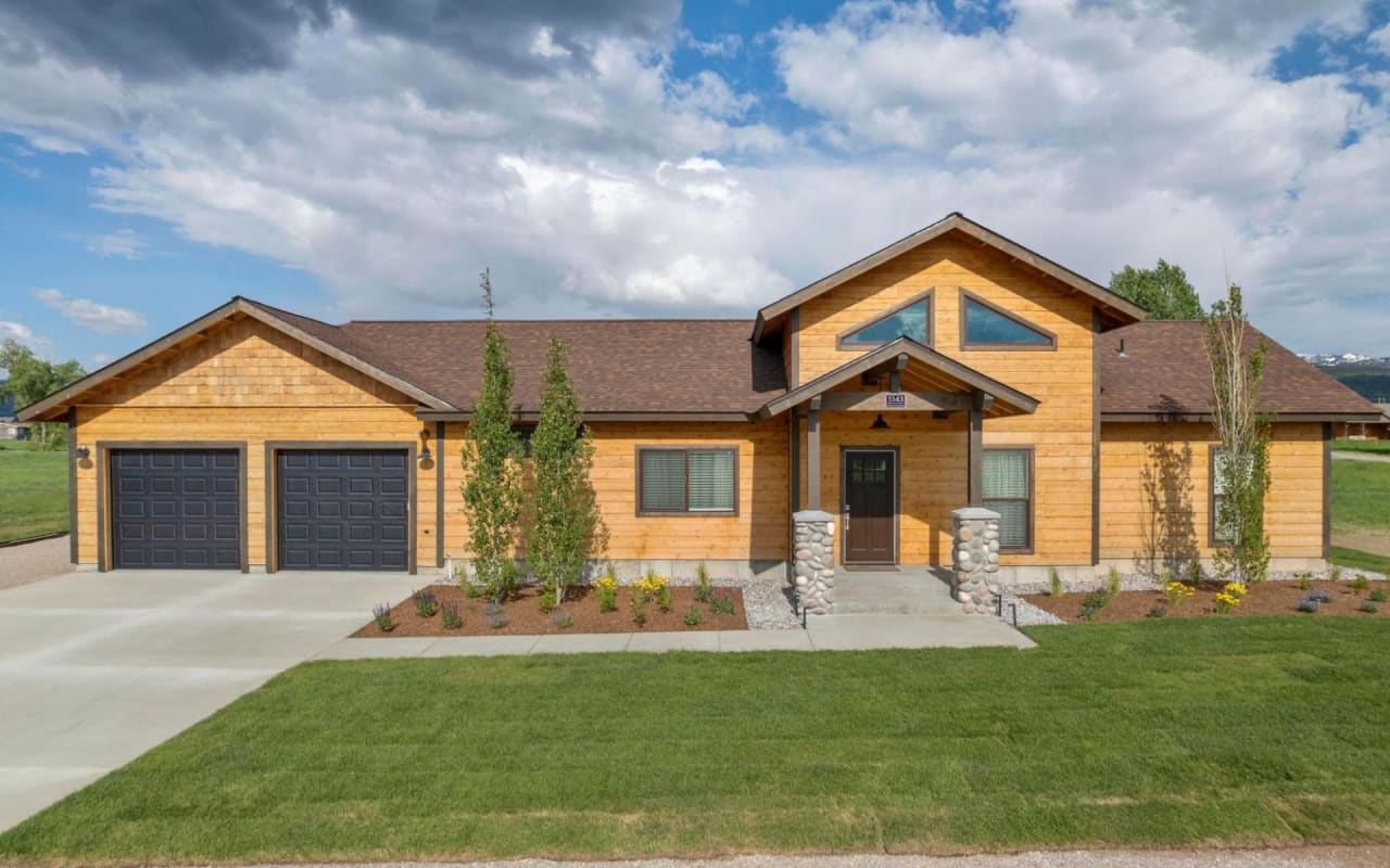 A large, two-story wooden house with a garage, dormer windows, covered porch, front yard, and gravel driveway.