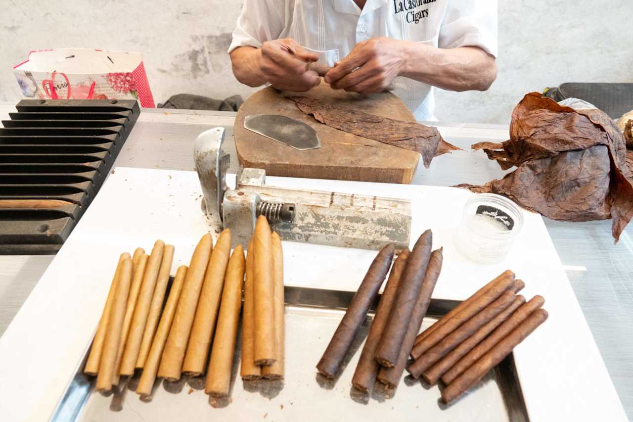 A cigar maker hand-rolls cigars beside tobacco leaves, tools, and finished cigars.