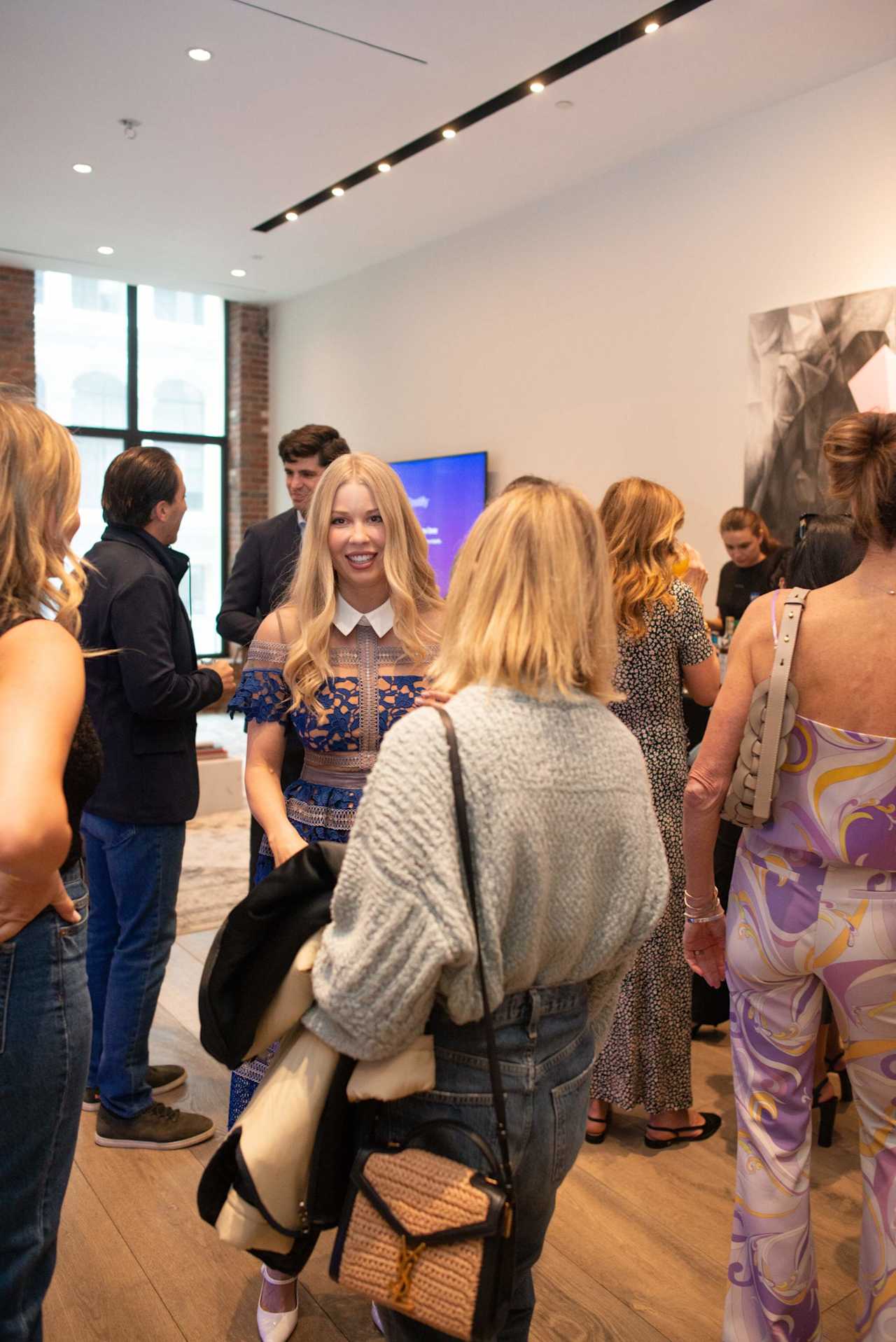 Guests mingle at a real estate event as a woman in a blue lace dress talks with attendees.