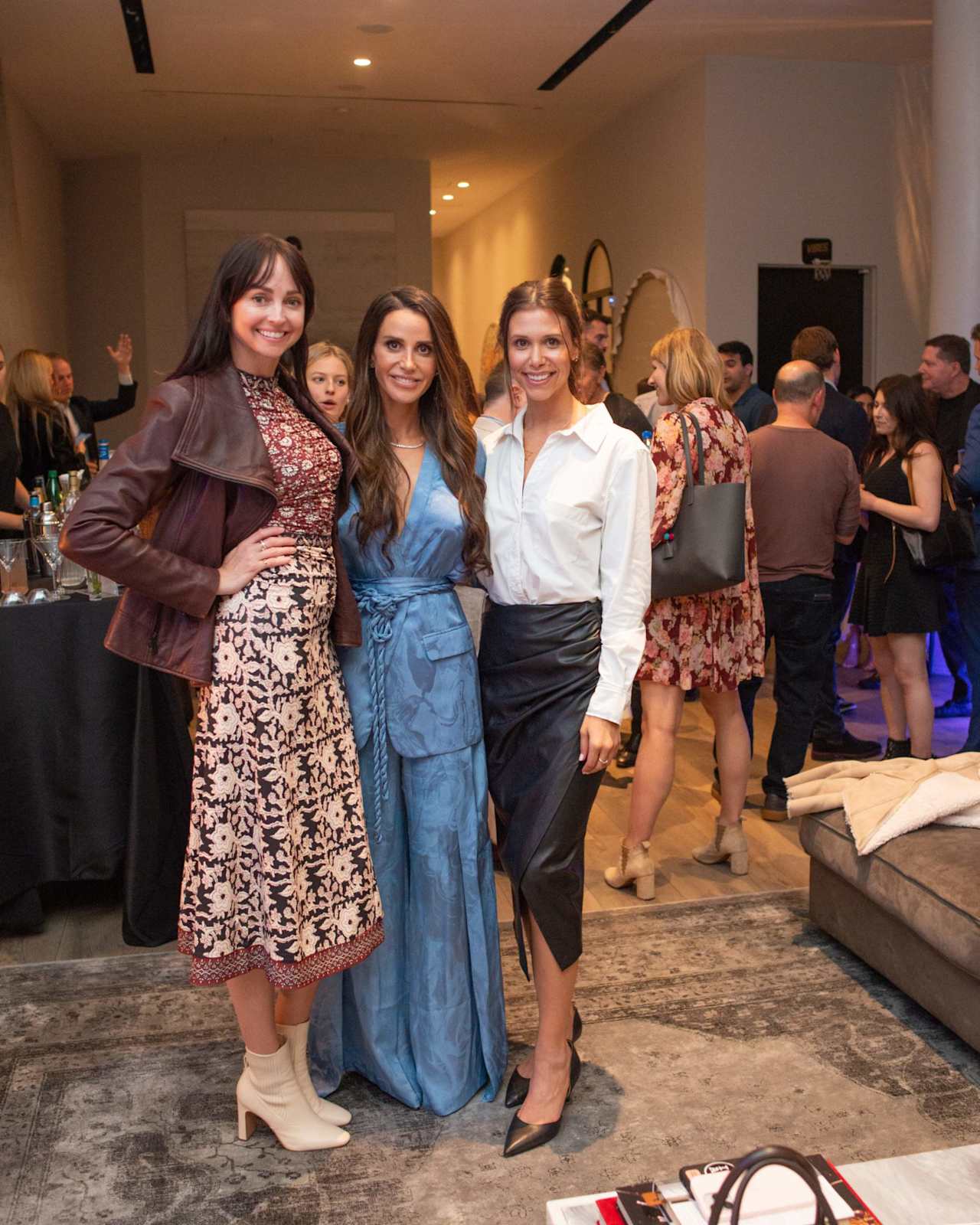 Three women pose together at an indoor gathering, wearing a blue dress, a white blouse and skirt, and a patterned dress.