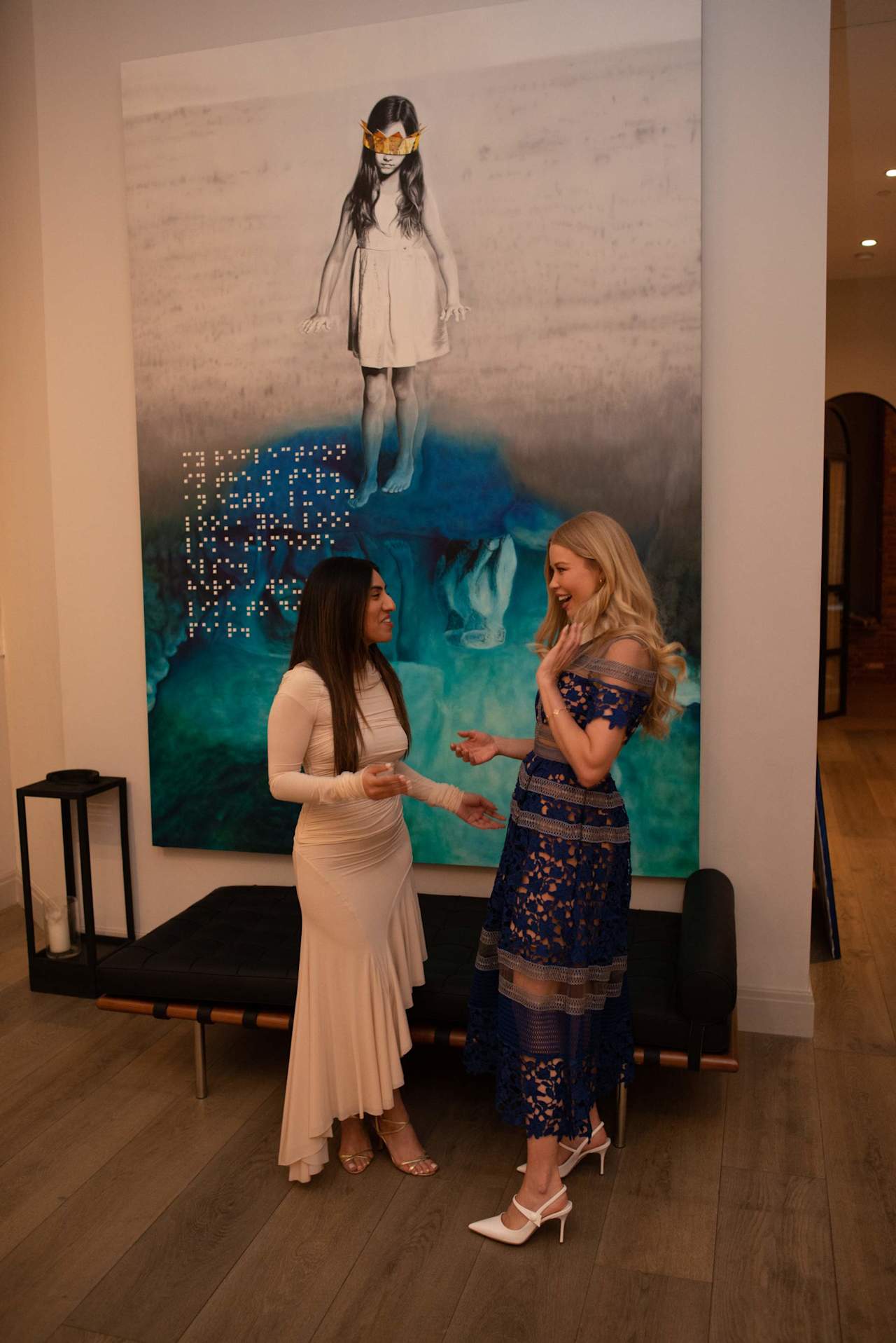 Two women in evening dresses talk in front of a large painting of a girl wearing a crown.