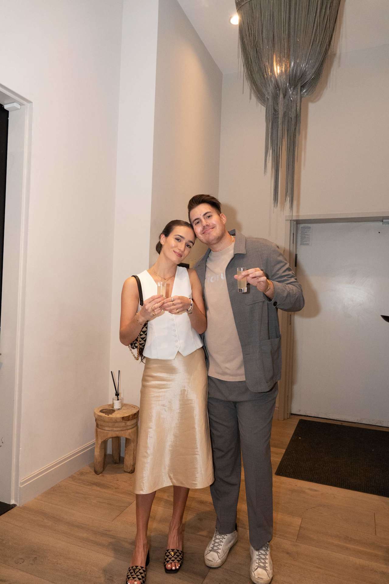 A couple pose with drinks beneath a hanging light at an indoor gathering.