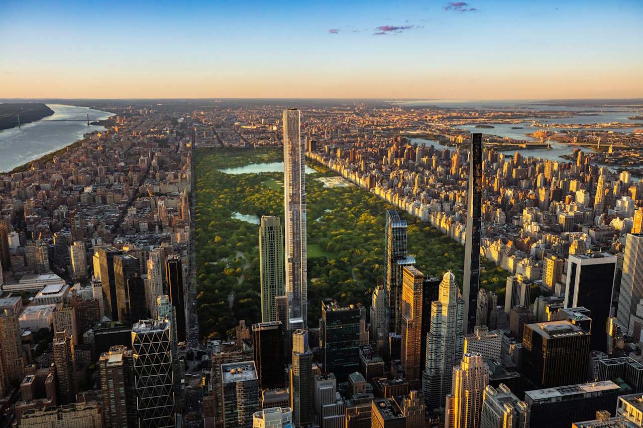Aerial view of a city skyline featuring tall buildings and a large green park, with rivers in the background under a clear sky.