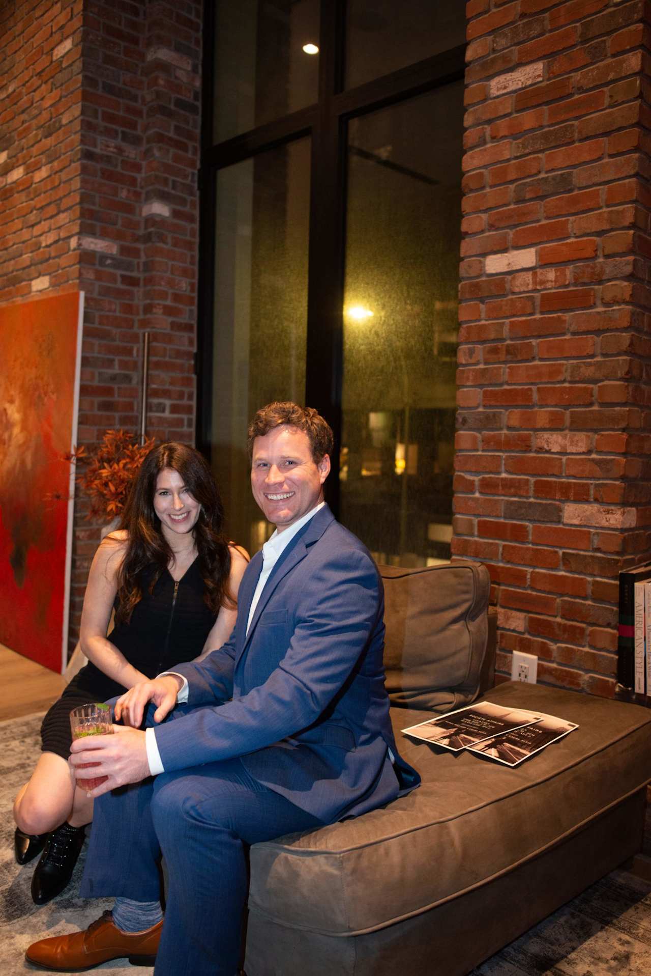Two adults seated together on a sofa in a brick-walled lounge; the man wears a blue suit.