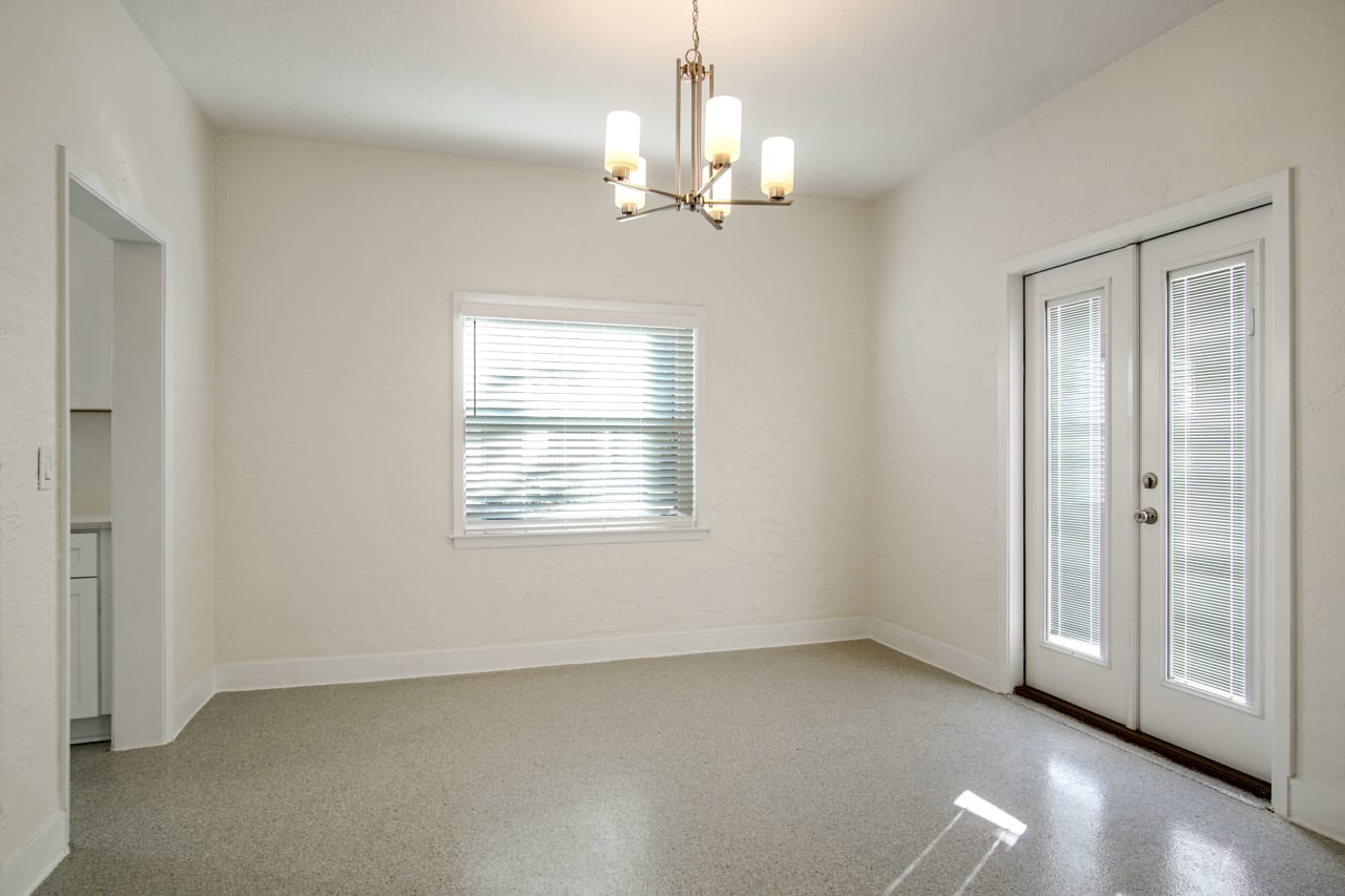 A dining area with a chandelier, a window, and a door leading outside. The room has a clean, neutral color palette and tiled flooring.