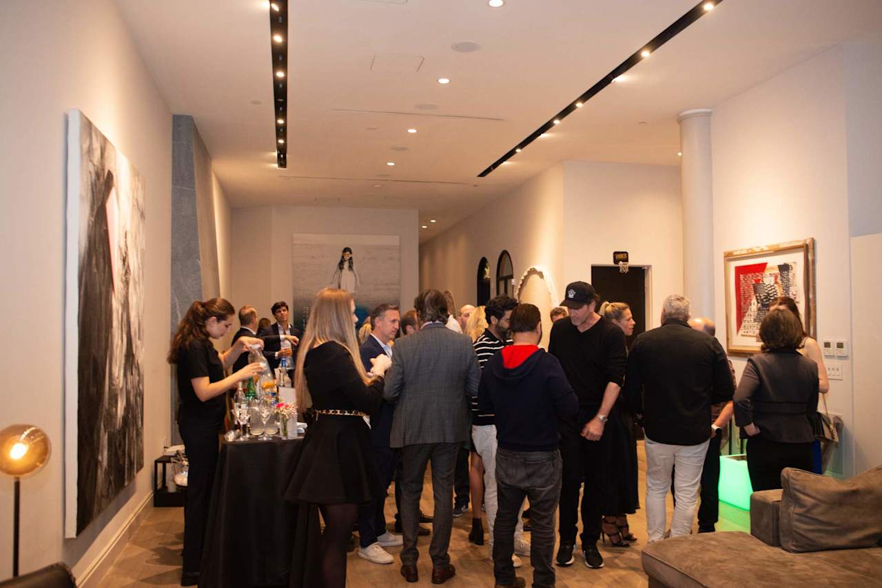 Guests mingle at a gallery gathering beside a table of drinks and framed artwork.