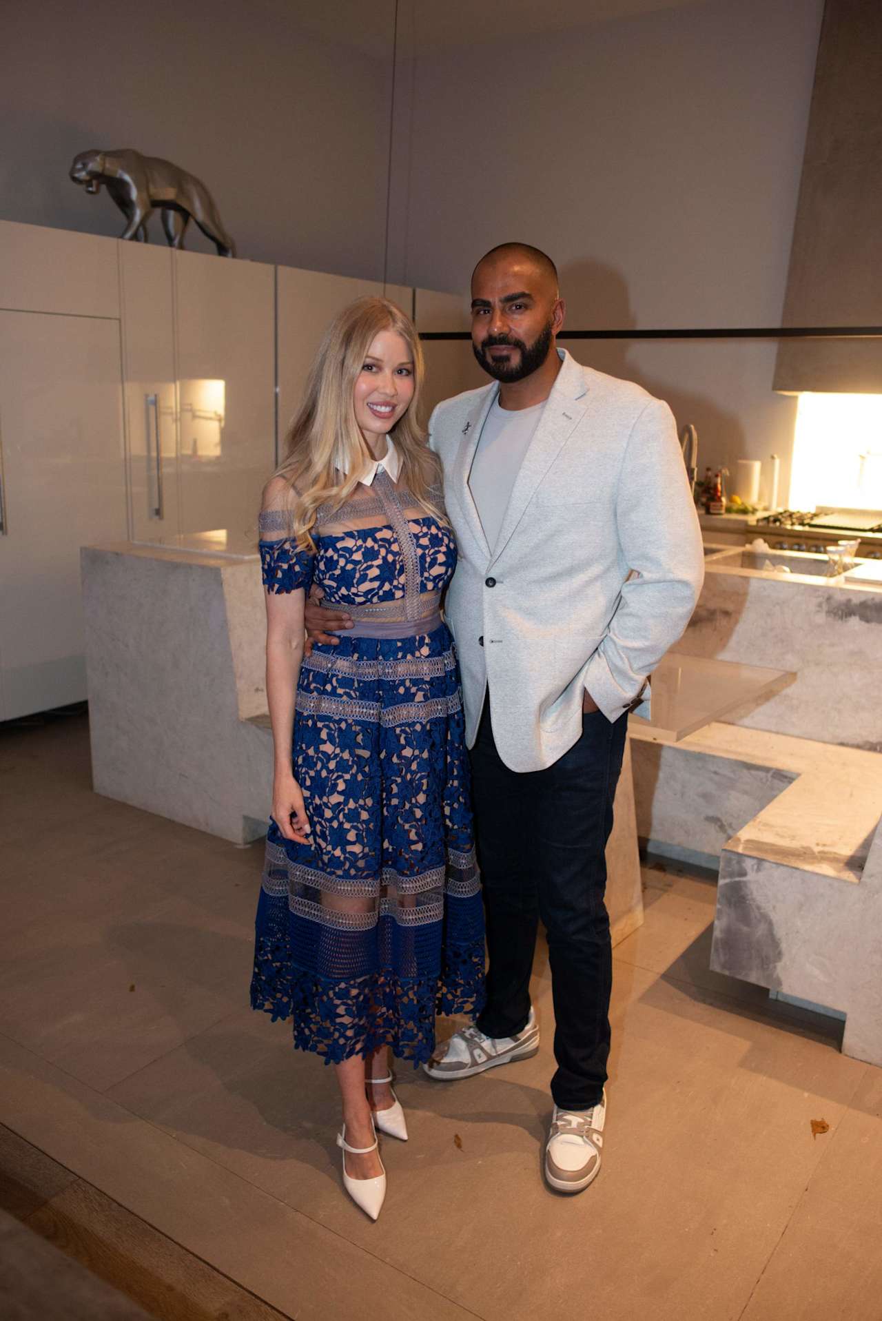 Two people pose in a modern kitchen, wearing blue lace and a light blazer.