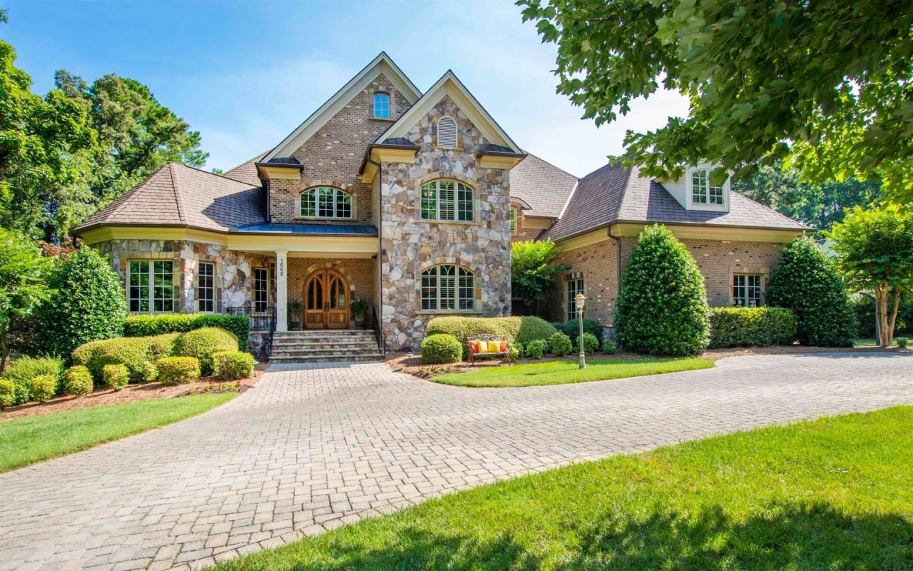 A two-story Tudor-style house on a large lot with a brick driveway, a half-timbered exterior, a slate roof, and a front yard.