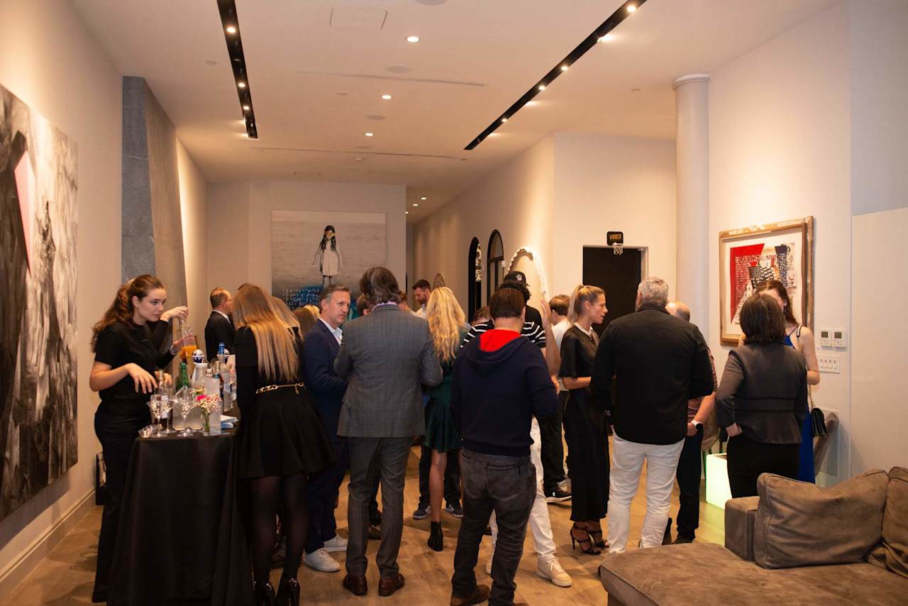 Guests mingle at a reception beside a drinks table and framed artwork.