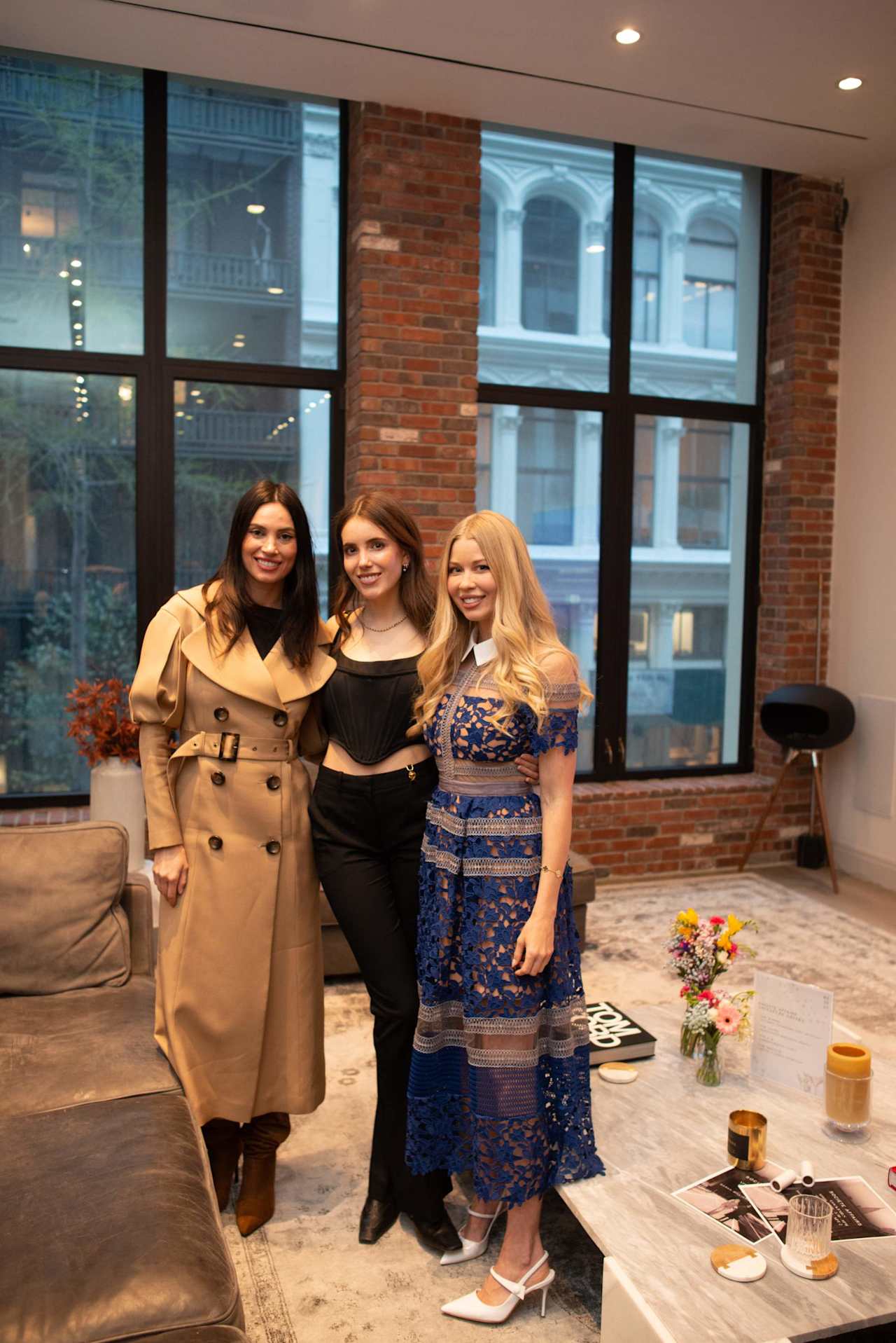Three women pose together in a brick-walled office lounge.