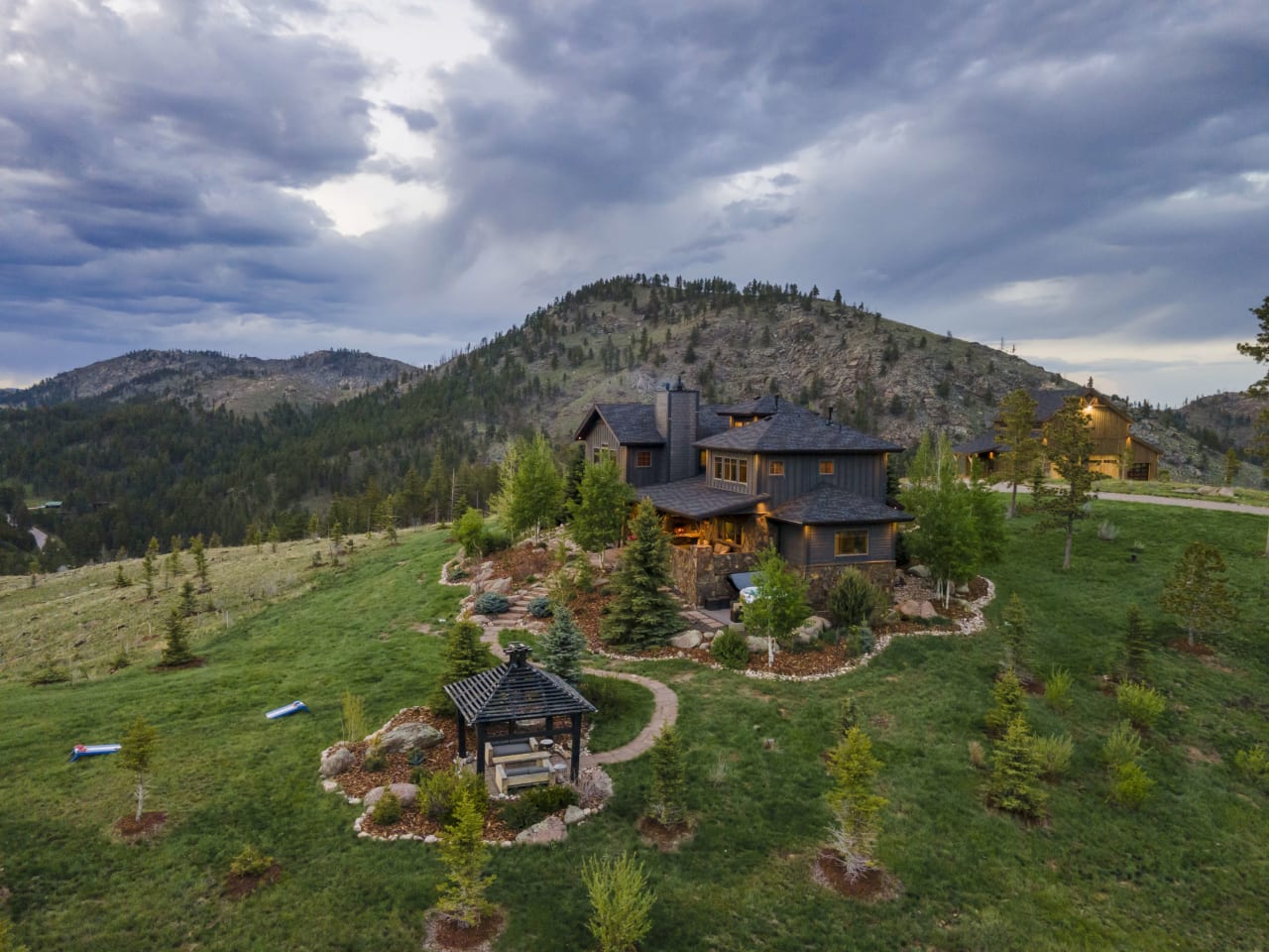 A large, rustic house sits on top of a hill, surrounded by evergreen trees and grass.