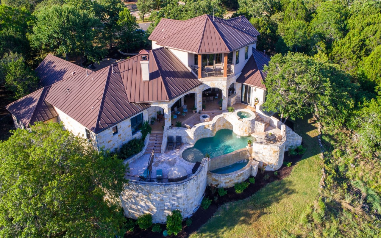 An aerial view of a large house surrounded by trees, with a swimming pool, patio, and a large stone wall.