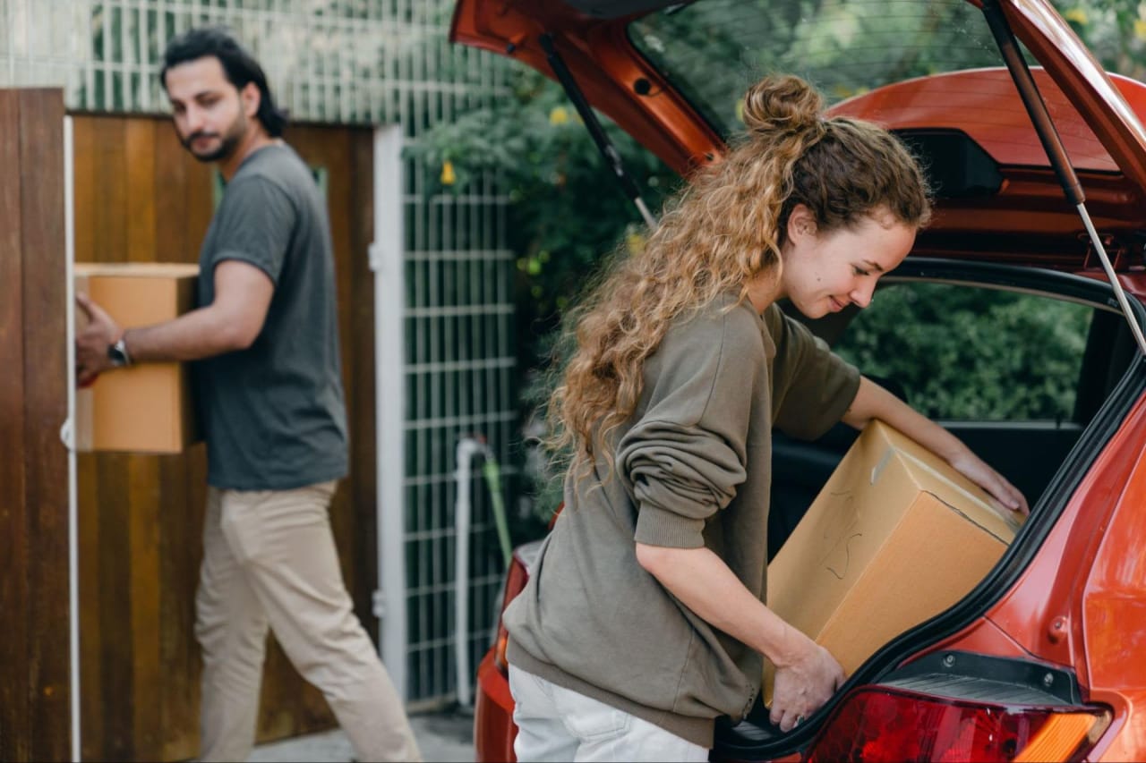 A woman unloading a box onto her car trunk, aided by a man during their relocation to a new home.